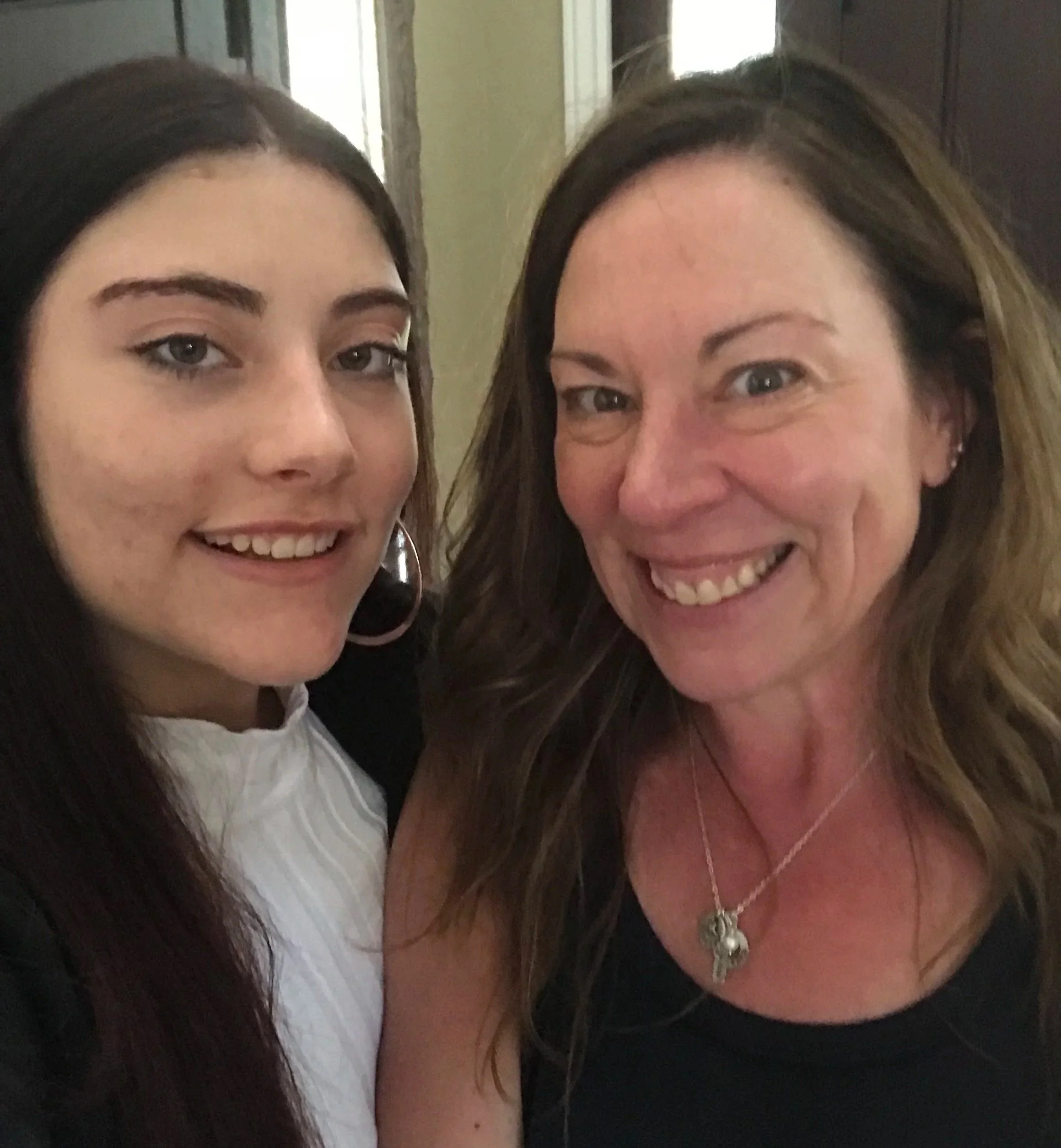 Two women (Lindsey and her mom) smiling for a selfie indoors, one with long dark hair and the other with long light brown hair, both wearing black tops.