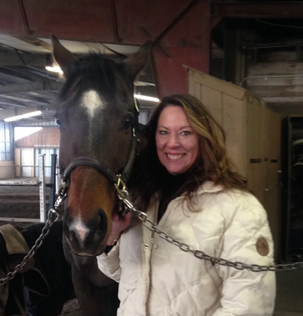 Woman (Lindsey's mom) with long brown hair smiling next to a horse inside a barn.