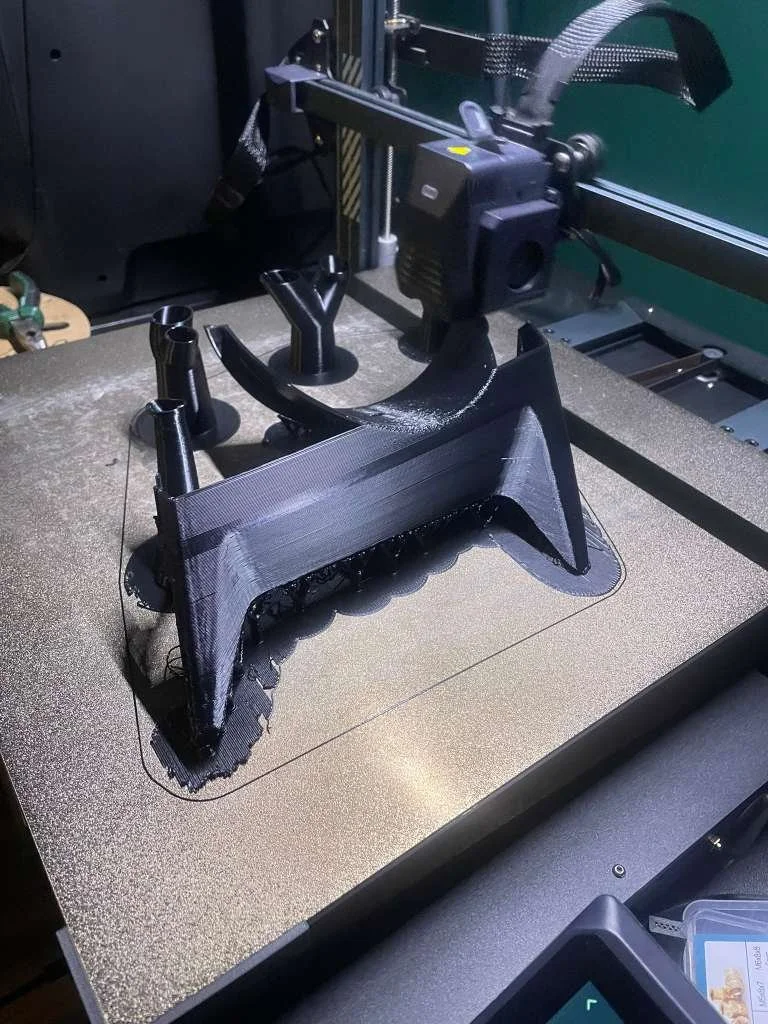 A black 3D printer creating a large, detailed black plastic object that looks like a toy or model, placed on a textured metallic surface.