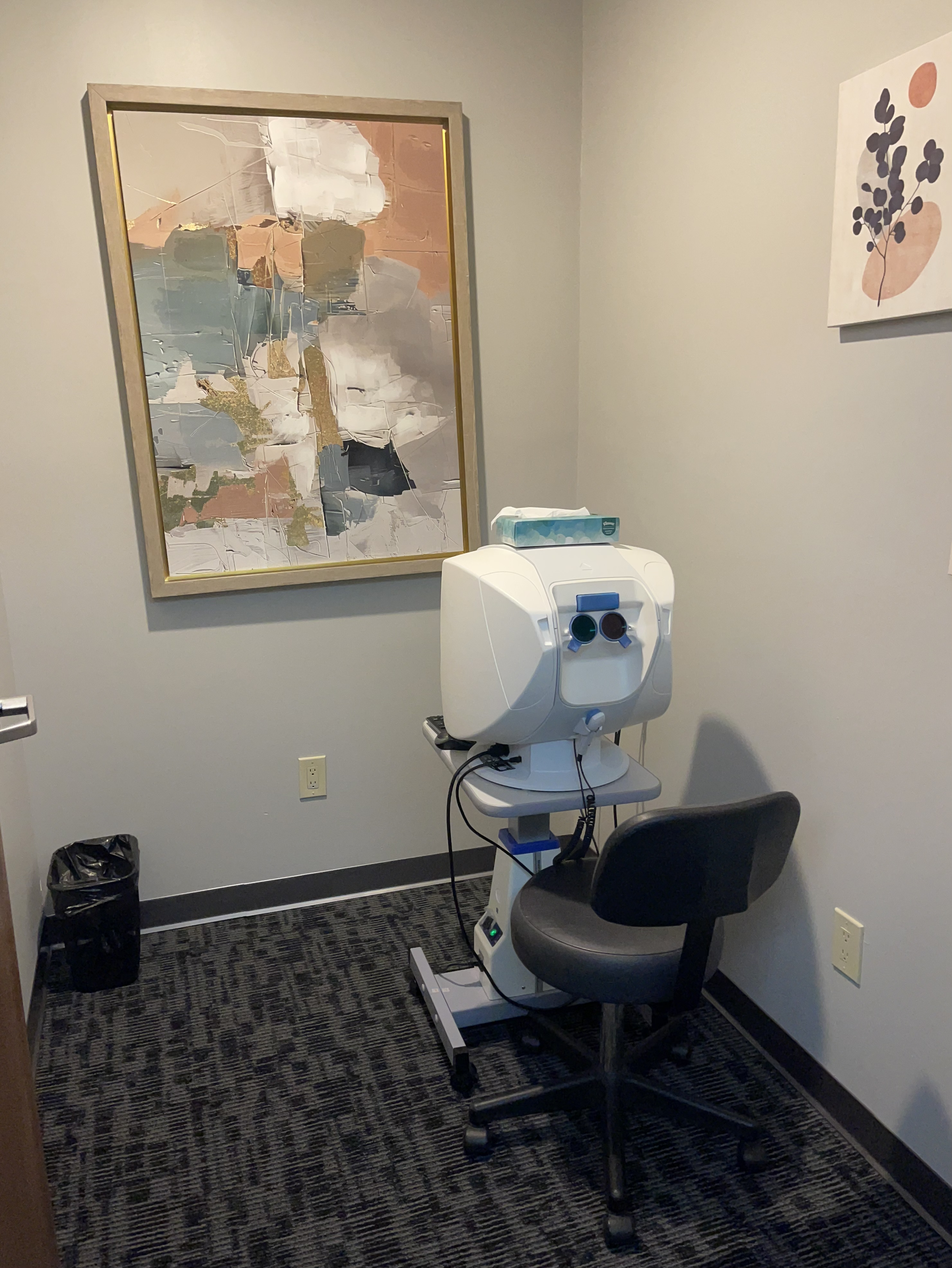 Optical coherence tomography (OCT) machine in a small examination room, with abstract artwork on the walls, a black chair, and a black trash can.