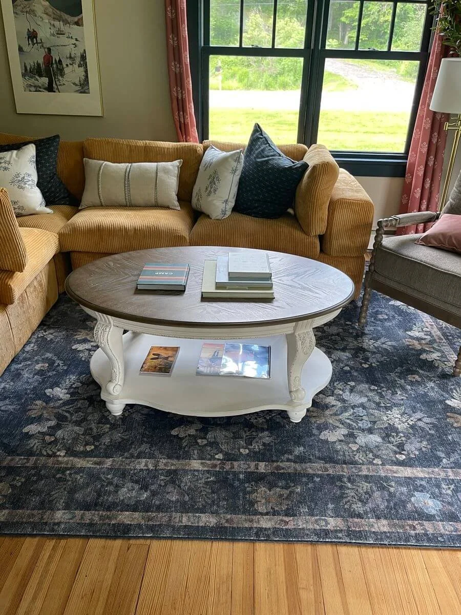 Living room with a yellow sofa, decorative pillows, a round wooden coffee table with books, a large window with pink curtains, and part of an armchair with a pillow, rug, and wooden floor.