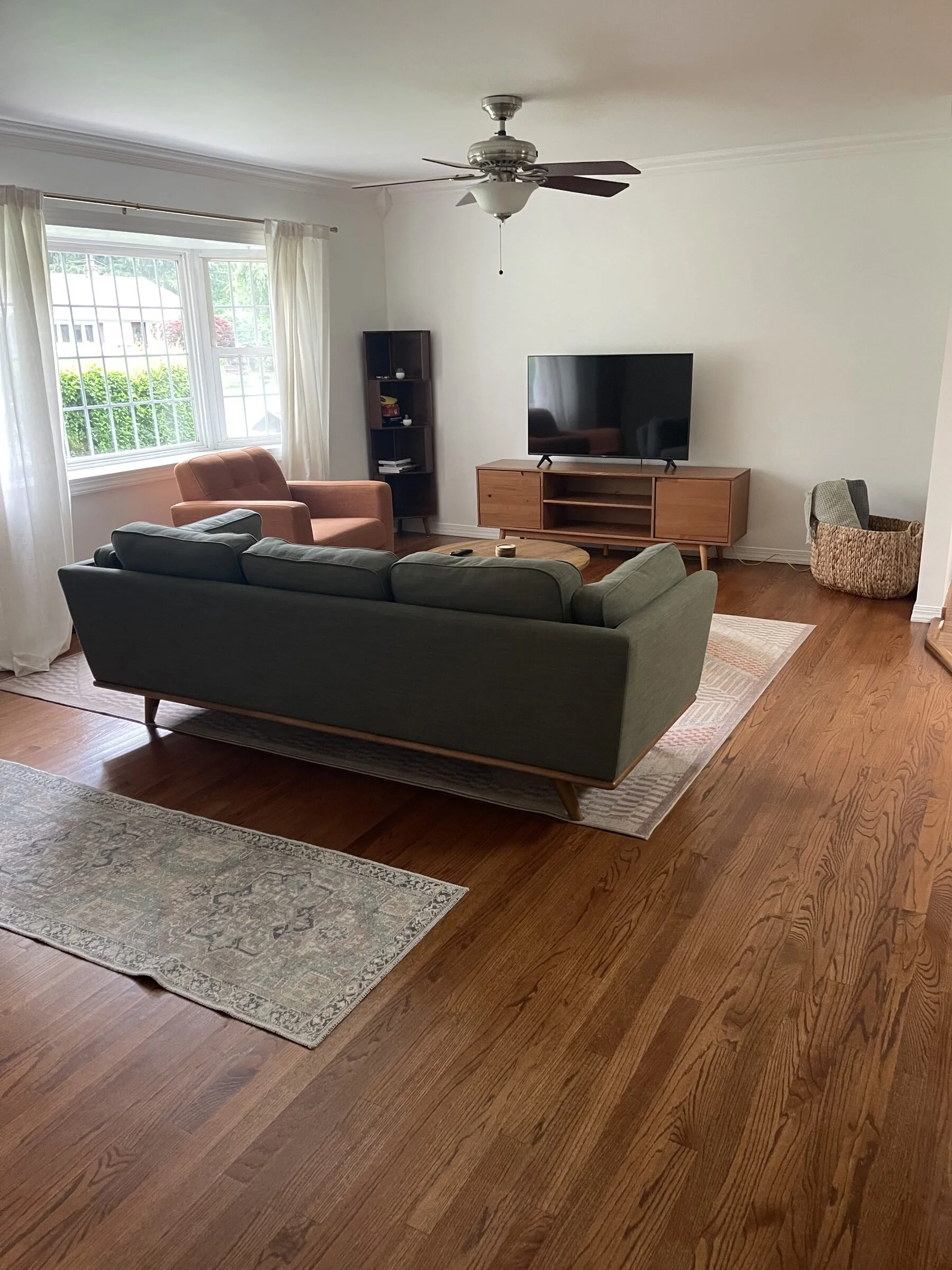 Living room with large window, green sofa, pink armchair, wooden television stand with a TV, small bookshelf, ceiling fan, area rugs, and hardwood flooring.
