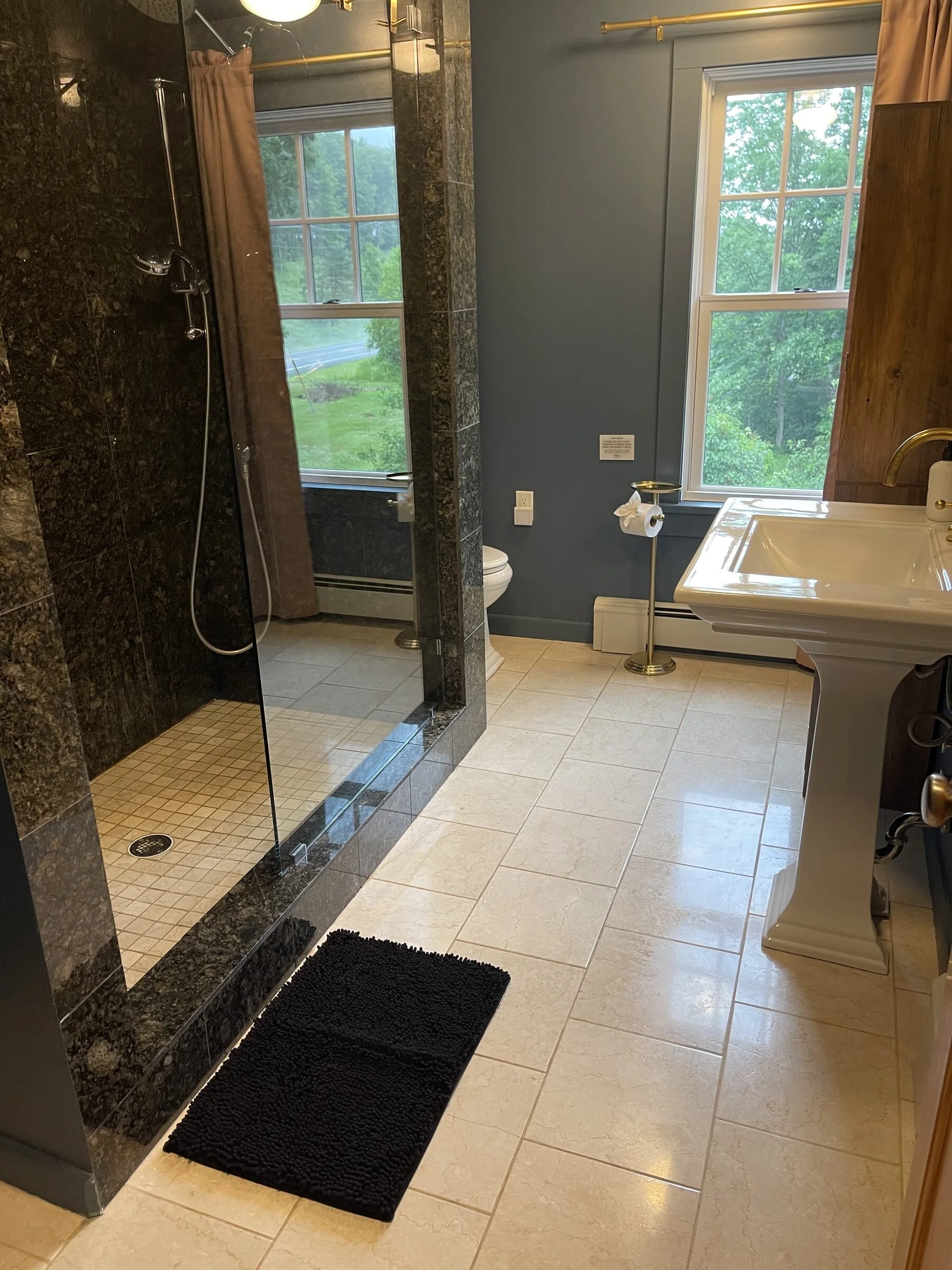 A bathroom with a walk-in shower on the left, featuring dark tile walls and a glass door. There are two windows letting in natural light, and a pedestal sink on the right. A black bathroom mat is on the beige tiled floor.