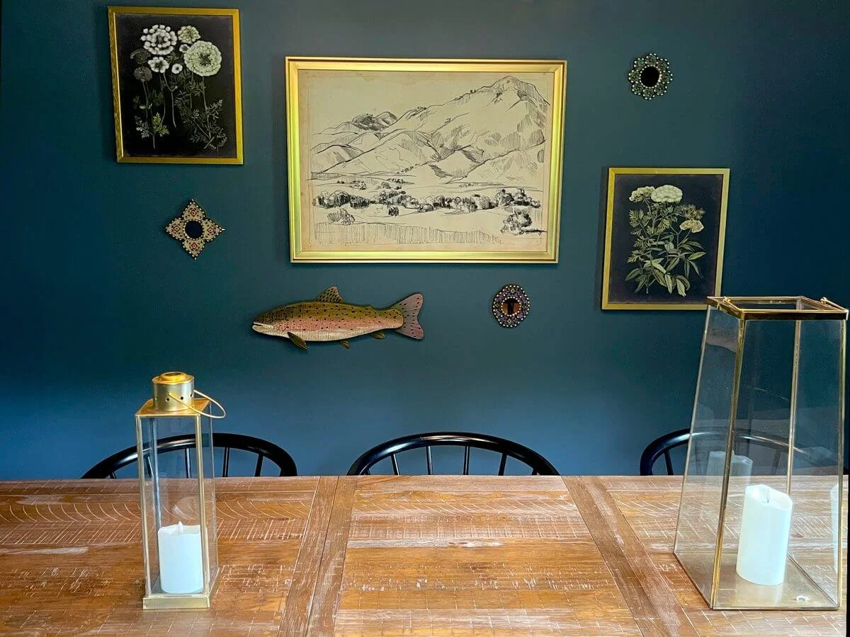 A wooden dining table with two gold-trimmed glass lanterns containing white candles, three black chairs, a blue wall with framed artwork including floral illustrations, a sketch of mountains and fields, and decorative mirrors, and a fish-shaped wall decor.