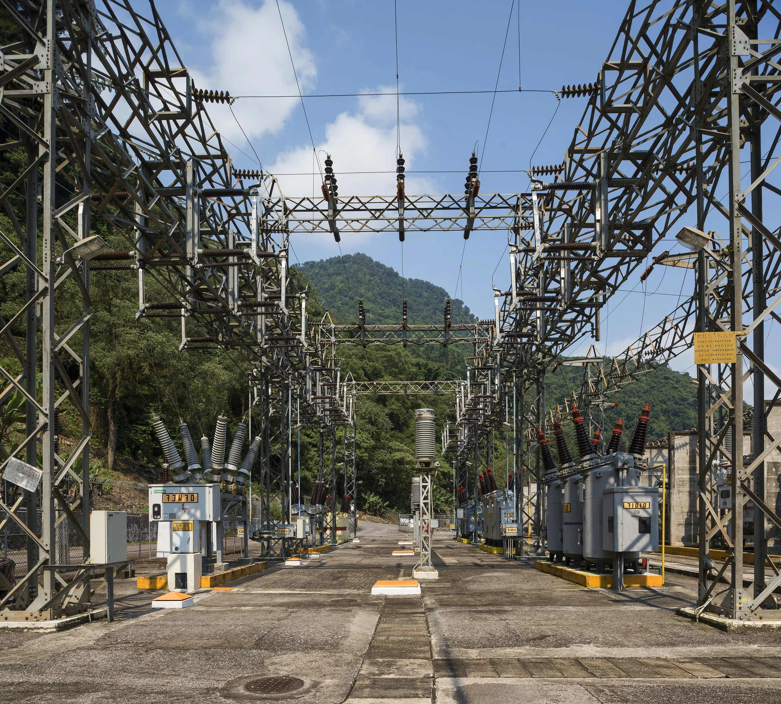 Vista aérea al atardecer de un embalse rodeado de montañas, bosques y una ciudad, con una represa visible en primer plano.