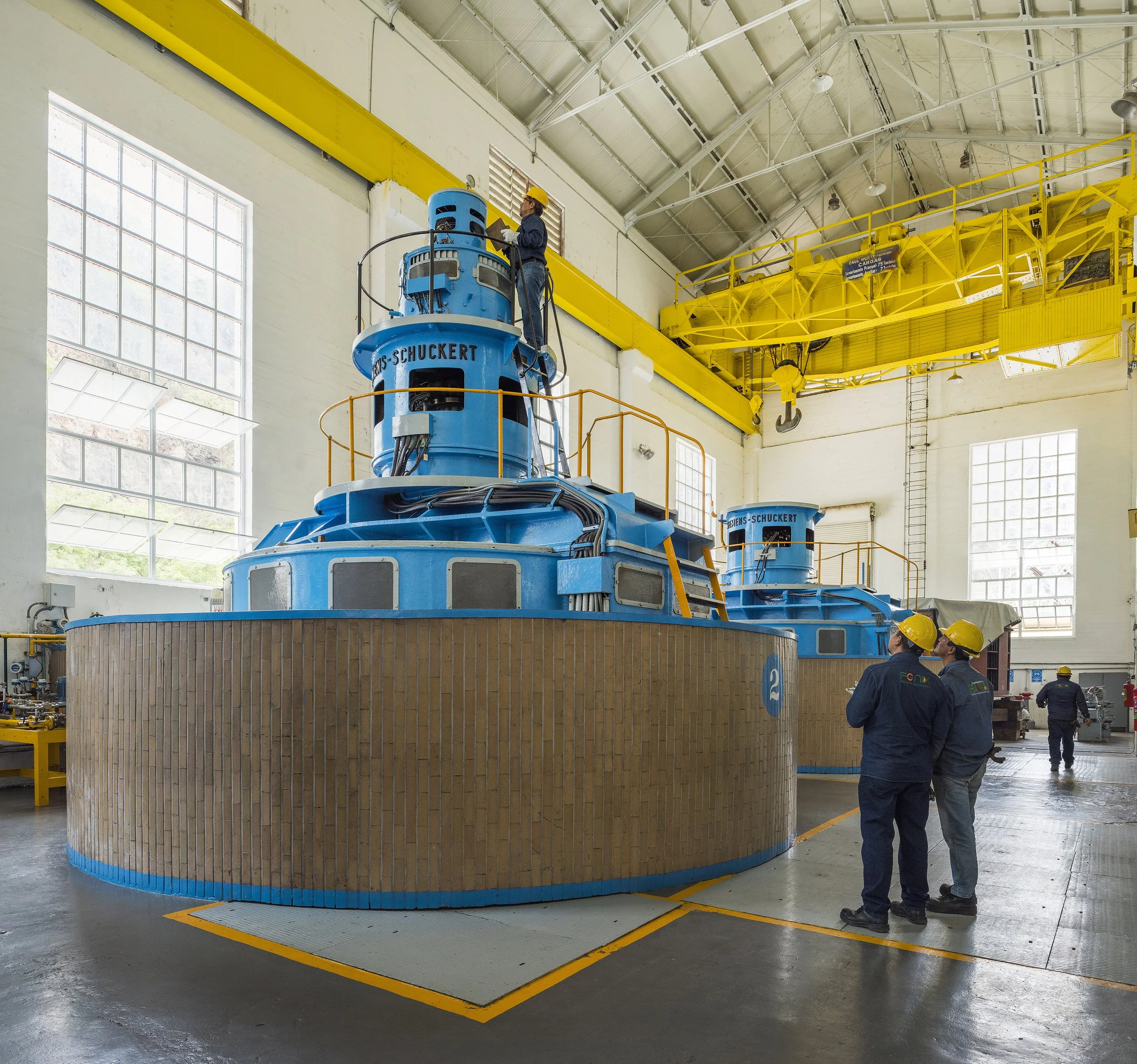Dos ingenieros eléctricos con cascos y equipo de protección inspeccionan una subestación eléctrica; uno de ellos señala hacia el horizonte mientras el otro sostiene un radio comunicador.