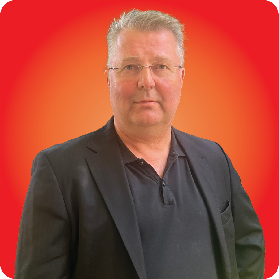 A middle-aged man with gray hair, wearing glasses, a black polo shirt, and a black blazer, standing against a red and orange gradient background.
