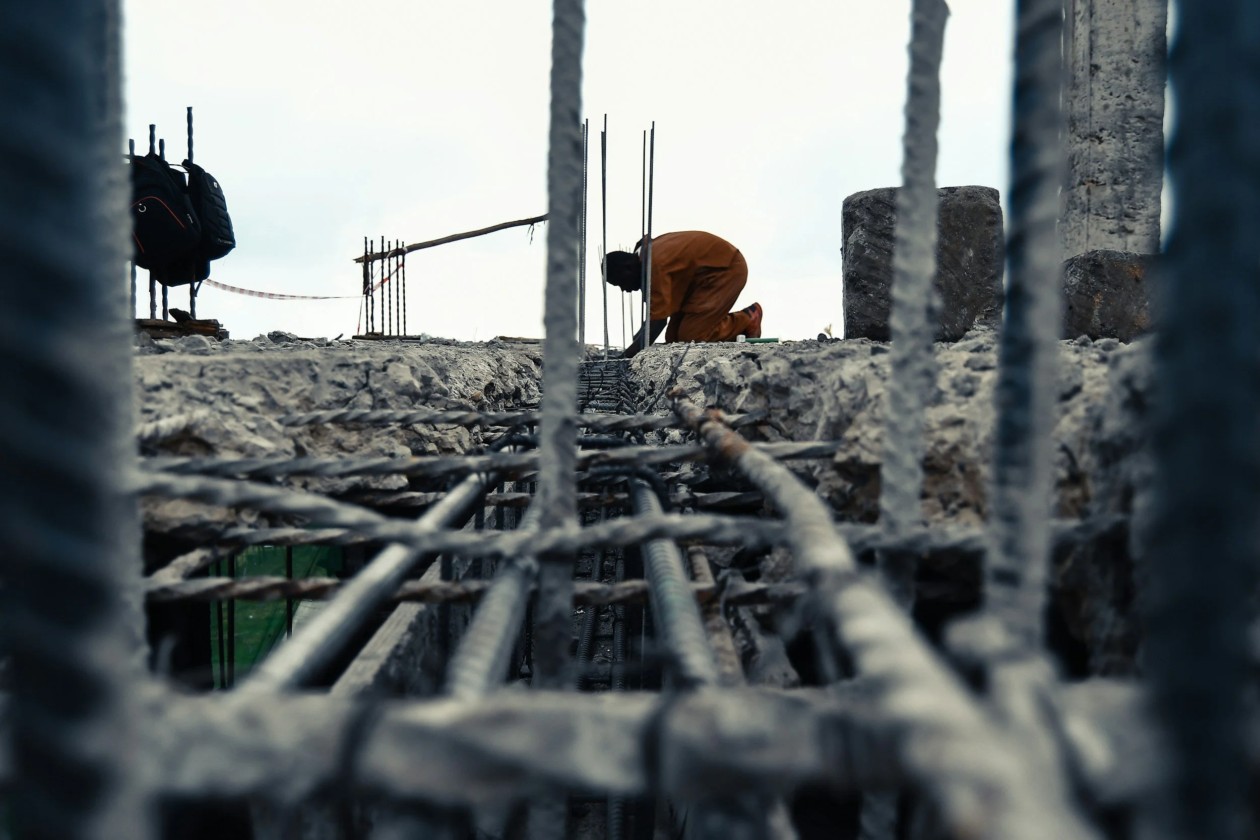 Construction worker kneeling on rebar reinforcement at a construction site, viewed through metal safety rails.