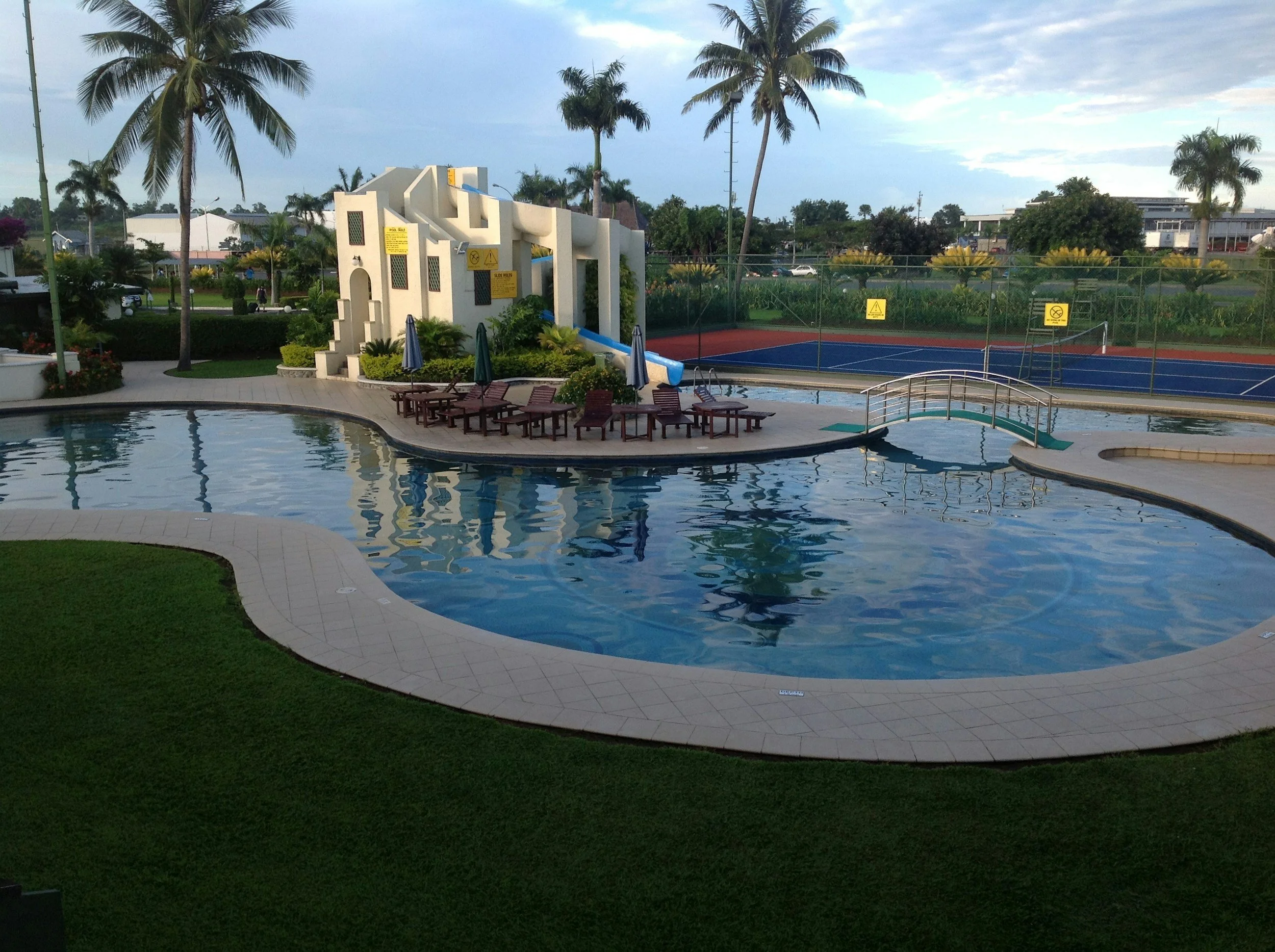 Pool area with lounge chairs and umbrellas, a water slide, and tennis courts in the background, surrounded by palm trees and greenery.