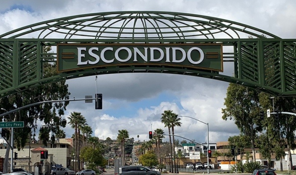 Sign that reads 'Escondido' under a decorative metal archway over a city street with palm trees and traffic lights.