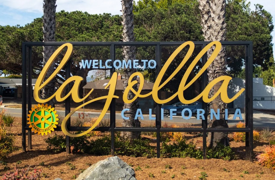 Welcome sign in La Jolla, California, with large yellow script reading 'La Jolla' and smaller blue text for 'Welcome to' and 'California,' and the Rotary International logo on the lower left.