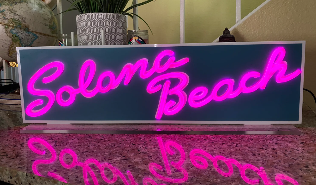 A neon sign that reads 'Solana Beach' in pink cursive lights, placed on a granite countertop with a reflection visible below.