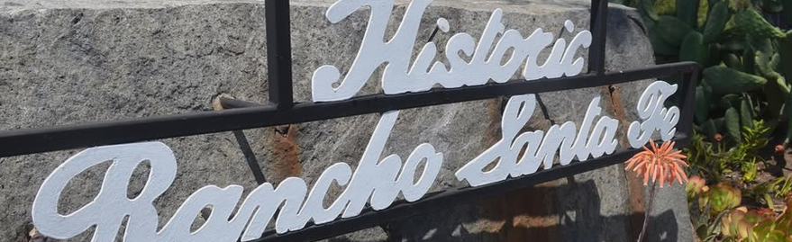 Metal sign reads 'Historic Rancho Santa Fe' in front of a stone wall, with plants nearby.