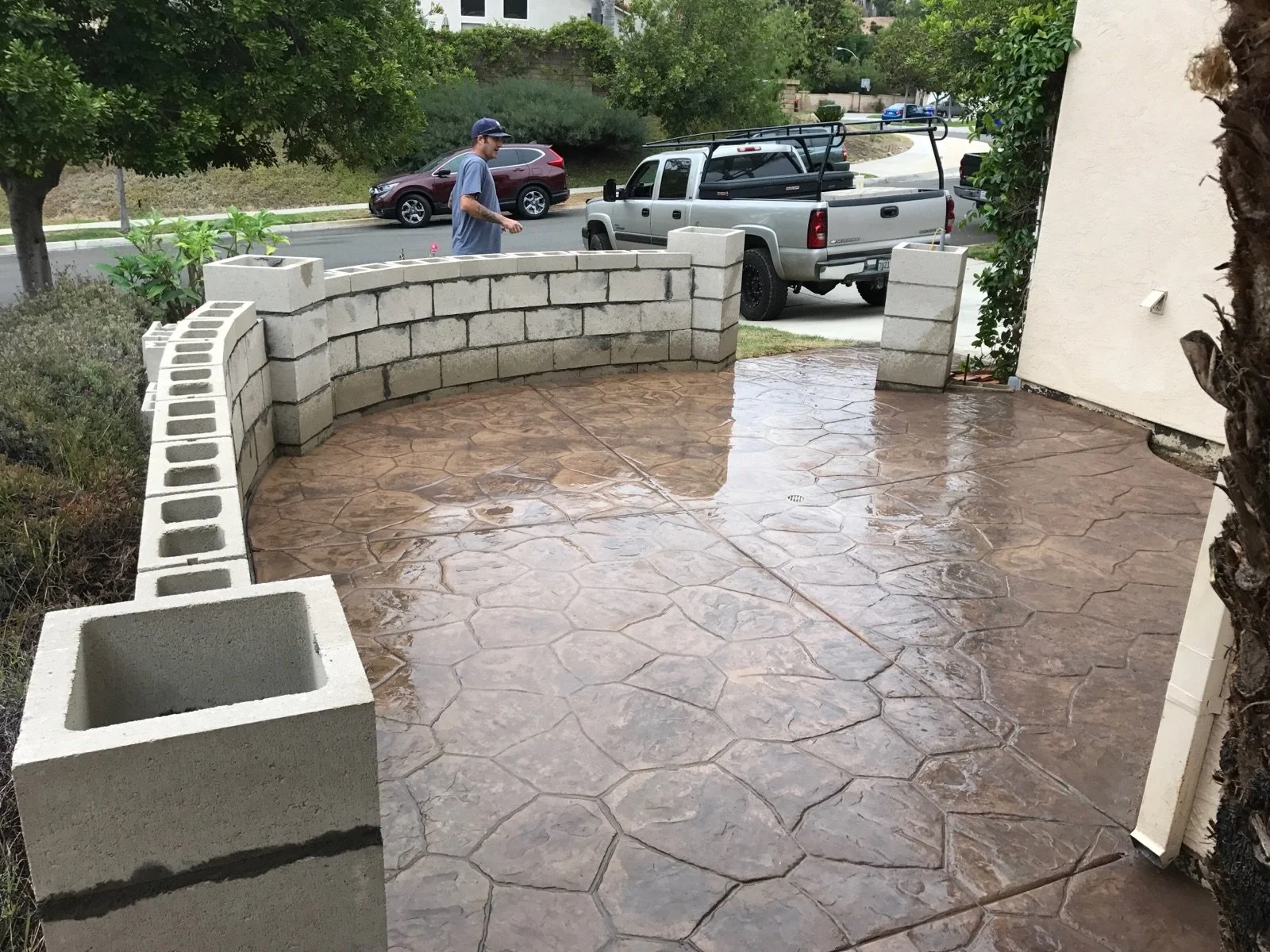 Wet concrete patio with decorative stamped pattern, enclosed by a short cinder block wall, with a person in a blue shirt and cap walking on the sidewalk outside, cars parked on the street, and greenery in the background.