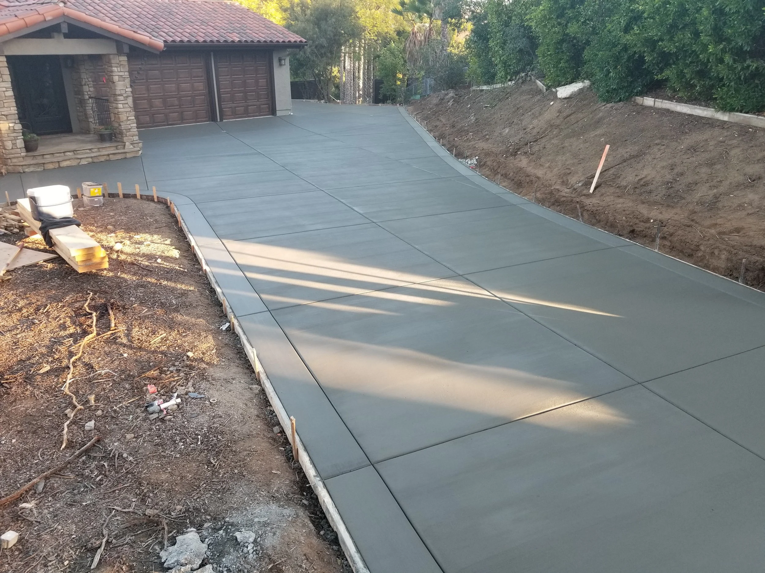 Newly poured concrete driveway in front of a house, with construction materials and dirt on the sides, and some trees and a fence in the background.