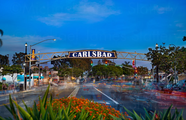 Street view in Carlsbad with a sign overhead, trees, parked cars, and a blue sky.