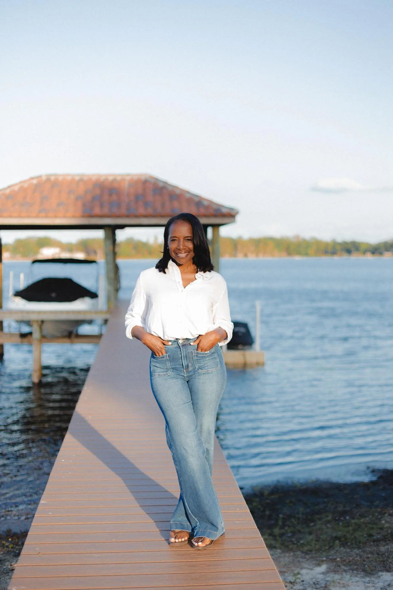 Woman standing on a dock by the water with a boat and a gazebo in the background, smiling at the camera.