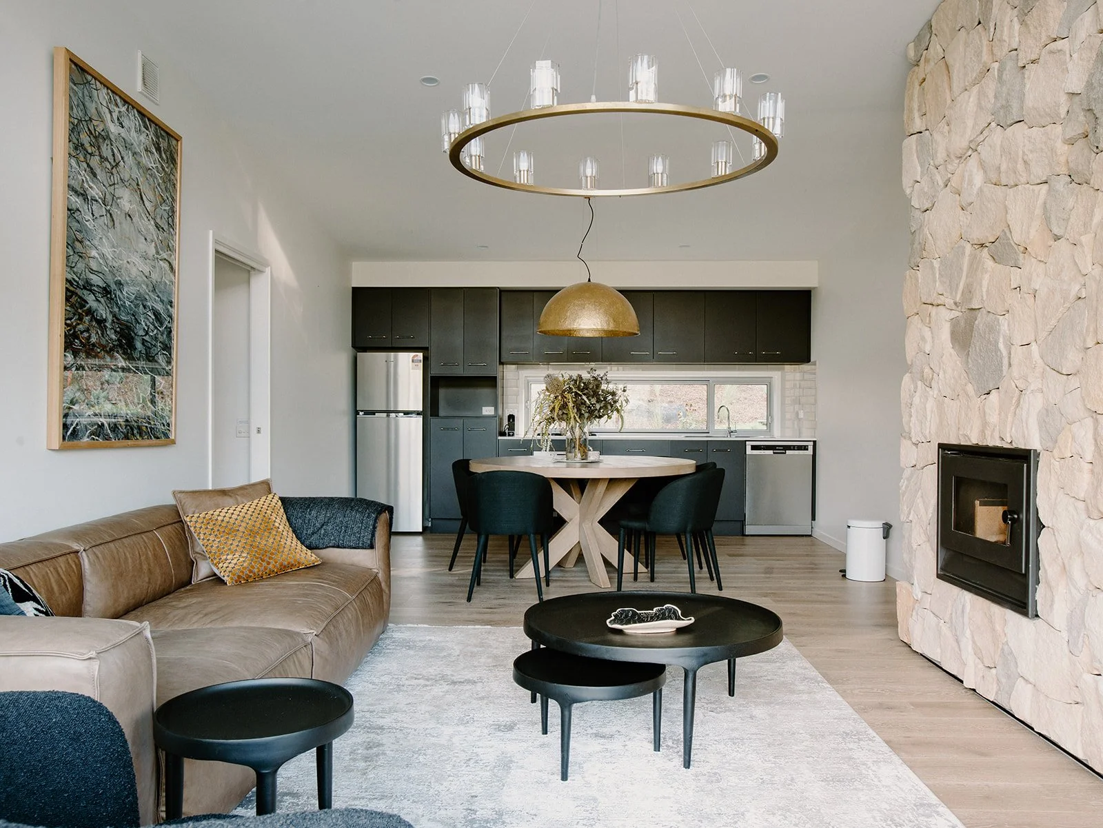 Open-plan living room and kitchen with modern decor, featuring a stone accent wall with a fireplace, black dining table with six chairs, black round side tables, beige leather sofa with patterned pillows, a large circular chandelier, and a kitchen wi