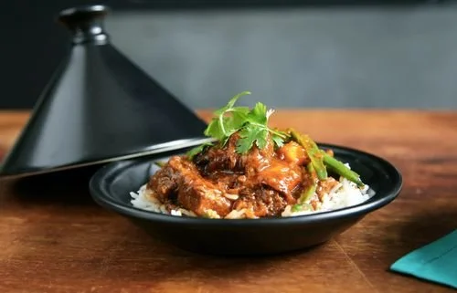 Beef stew served over rice with green herbs on top, in a black bowl with a black serving cover in the background.