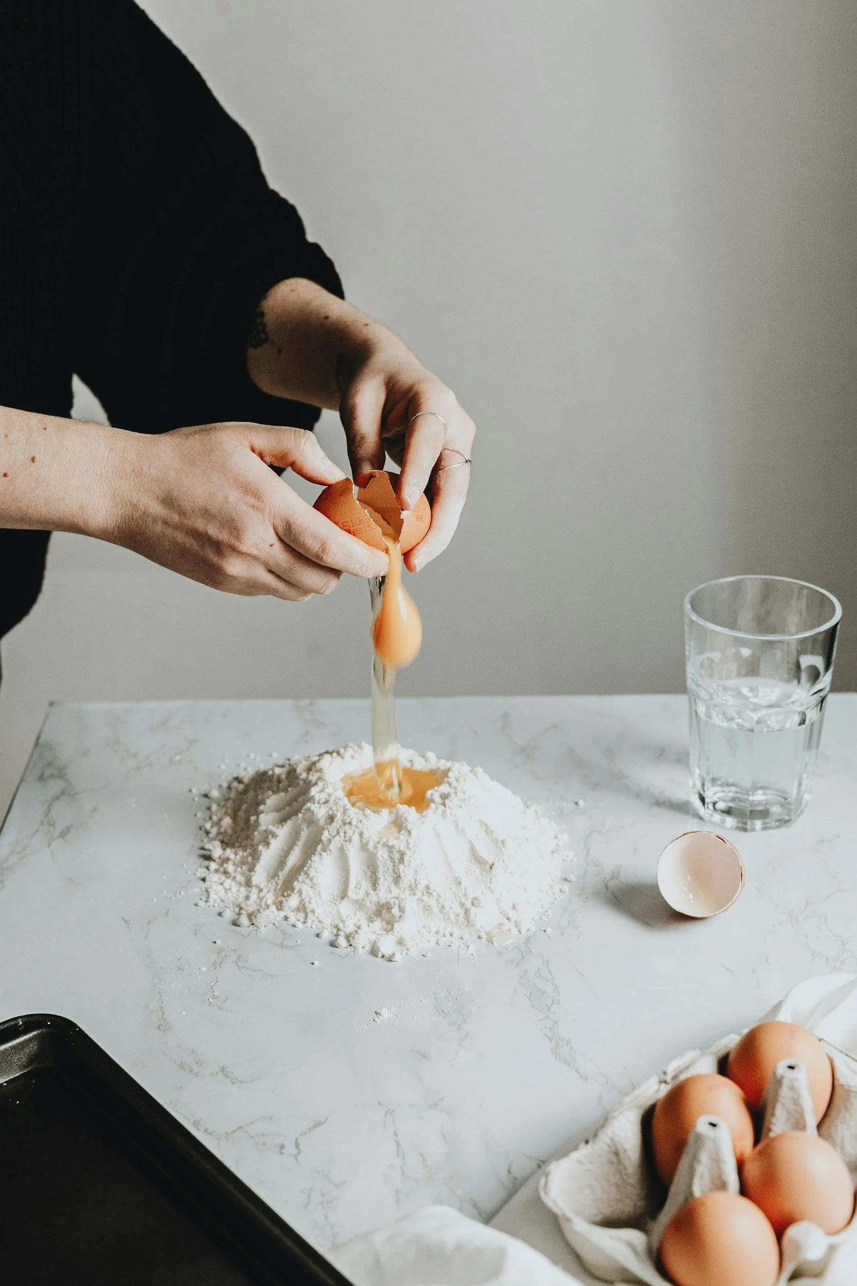 Person cracking an egg into a mound of flour on a marble surface with a glass of water nearby and an egg carton on the side.