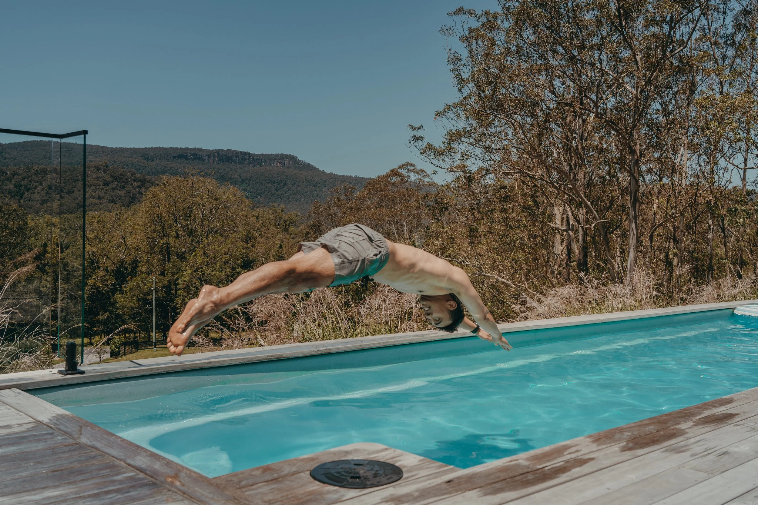 A man in swimming shorts diving headfirst into an outdoor pool from a wooden deck, with trees and mountains in the background on a clear day.
