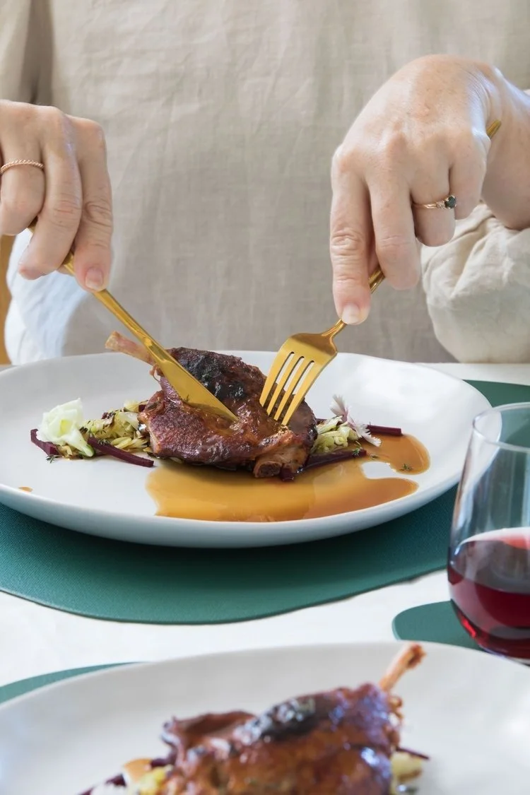 Person using a fork and knife to cut into a cooked piece of meat on a white plate, with some sauce and greens, in a dining setting with a glass of red wine nearby.