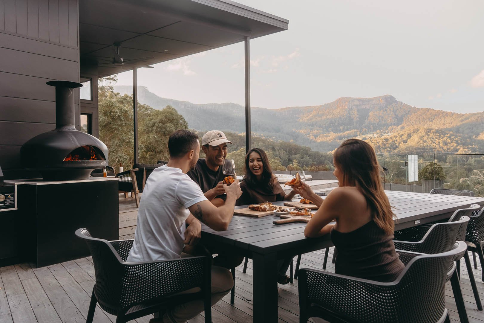Four friends enjoying pizza and drinks on an outdoor deck with scenic mountain views in the background.
