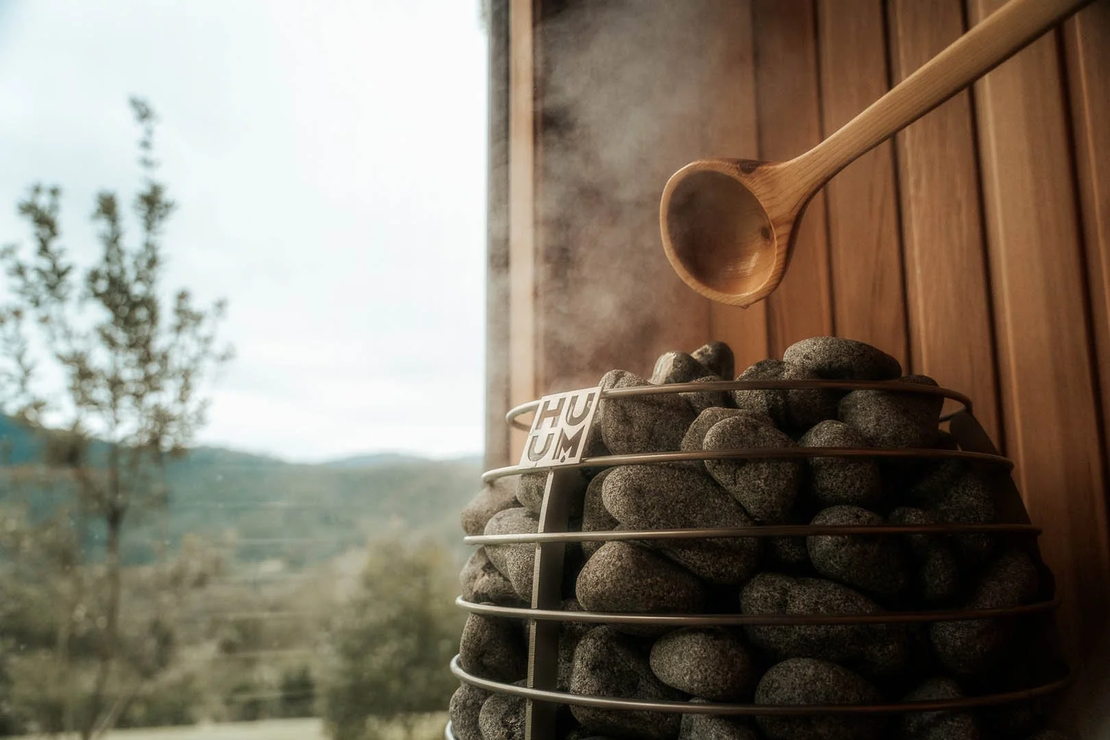 A wooden sauna scoop hanging above a sauna heater filled with rocks, with steam rising from the rocks, and a window showing an outdoor scene of trees and mountains.