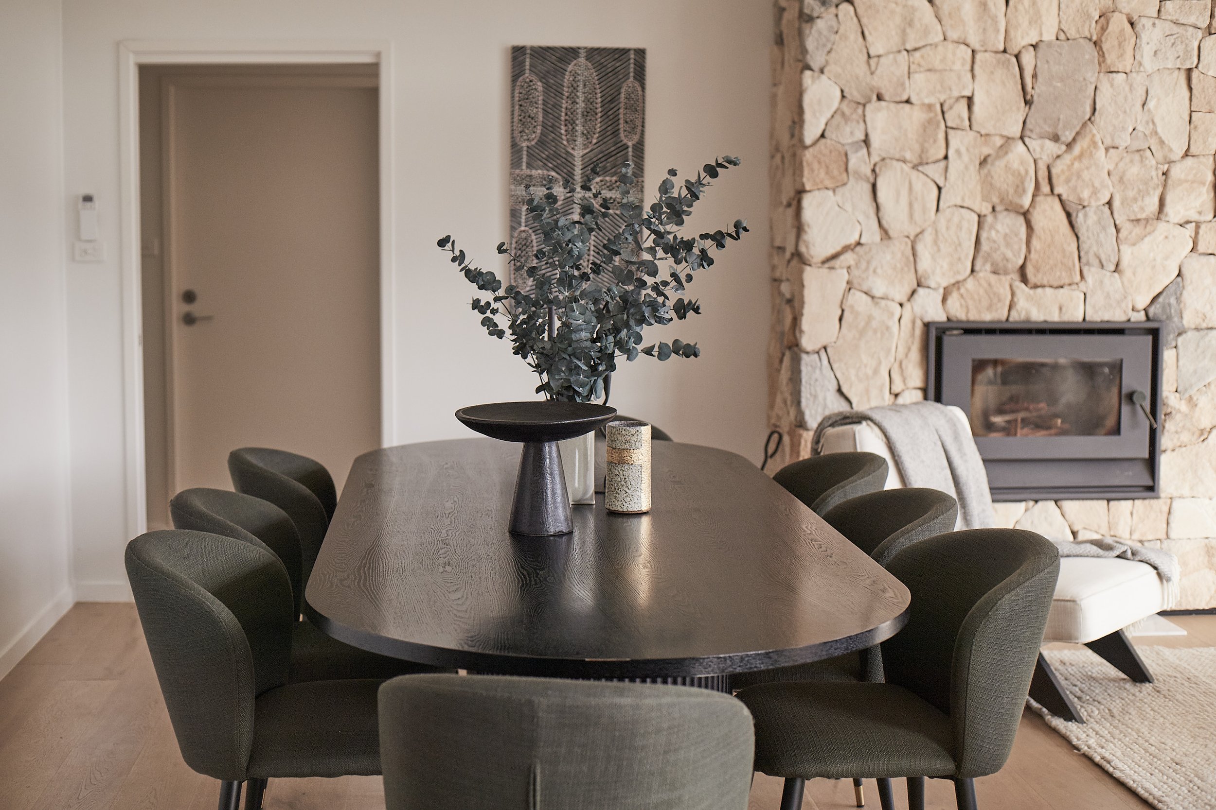 Living room with a dining table surrounded by eight black chairs, a stone fireplace, and a chair with a blanket, with decor on the wall and a vase with greenery on the table.