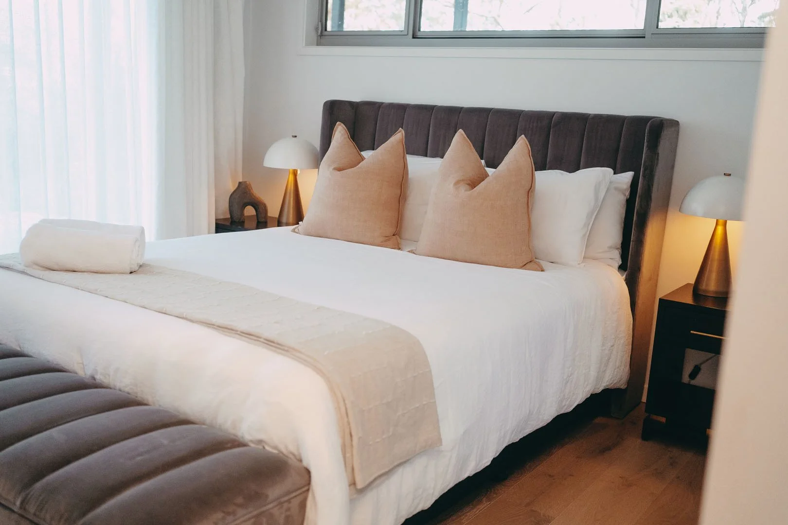 A neatly made bed with beige pillows, a rolled towel, and a cream-colored blanket, flanked by two table lamps with gold bases in a well-lit bedroom with large windows.