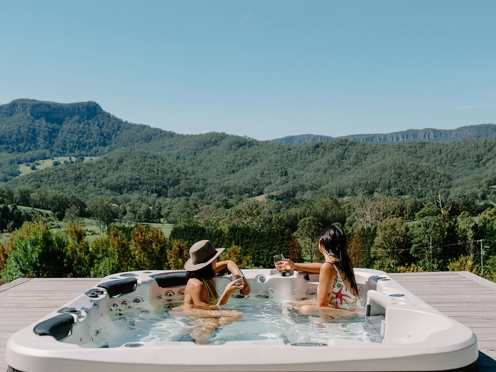 Two women relaxing in a hot tub on a wooden deck, drinking wine, with a scenic view of green hills and mountains under a clear blue sky.