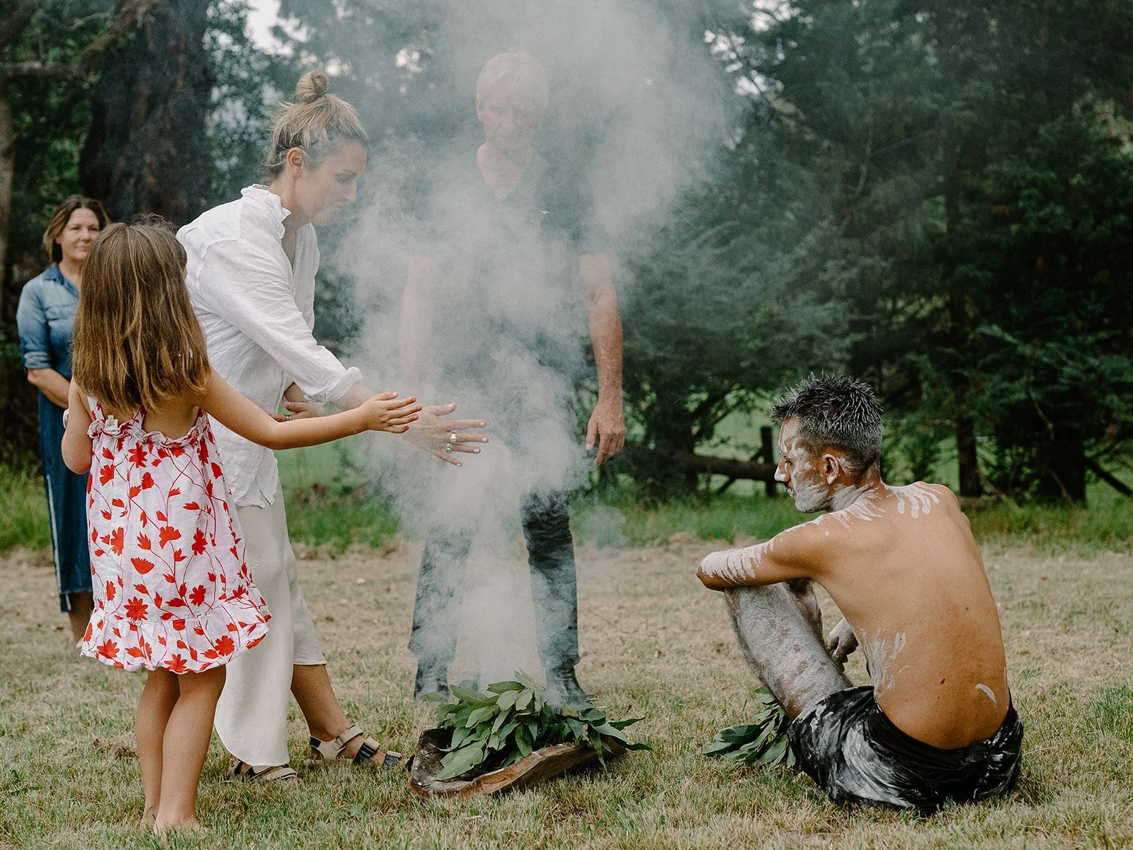 A group of people participating in a traditional Maori haka ceremony outdoors, with a man sitting on the ground covered in white paint or chalk, and others standing around him, some clapping and one woman reaching out towards him.