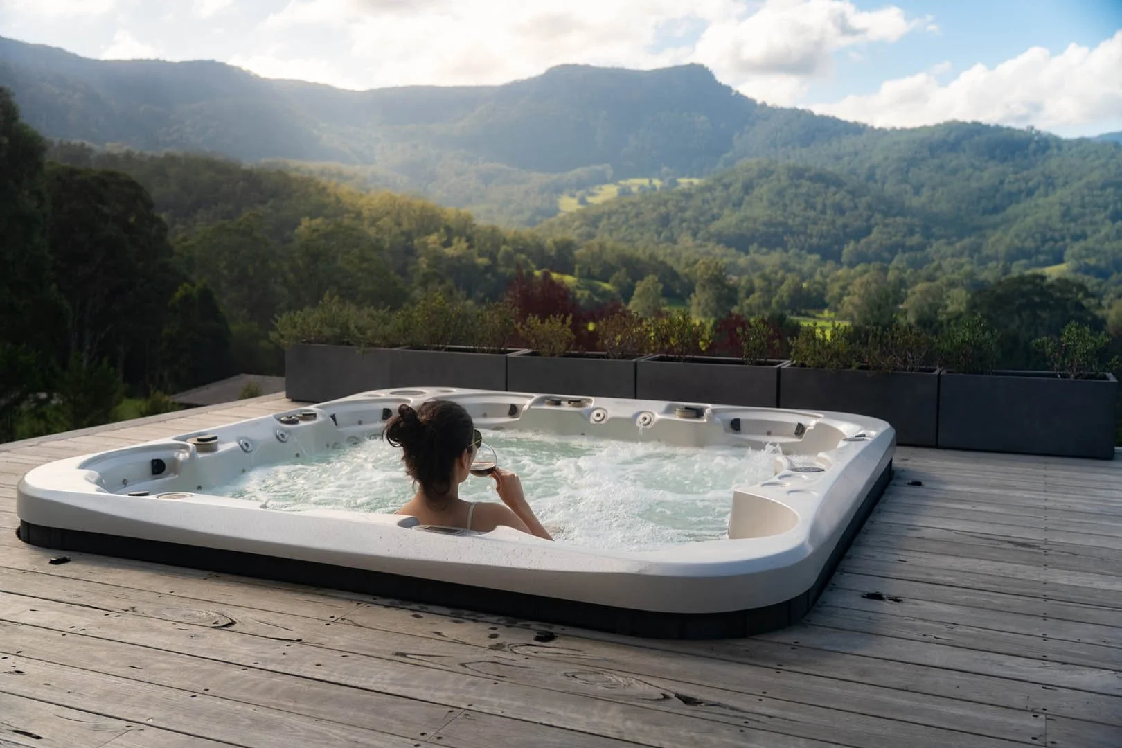 A woman in a hot tub on a wooden deck, overlooking a mountain landscape with greenery and clouds in the sky.