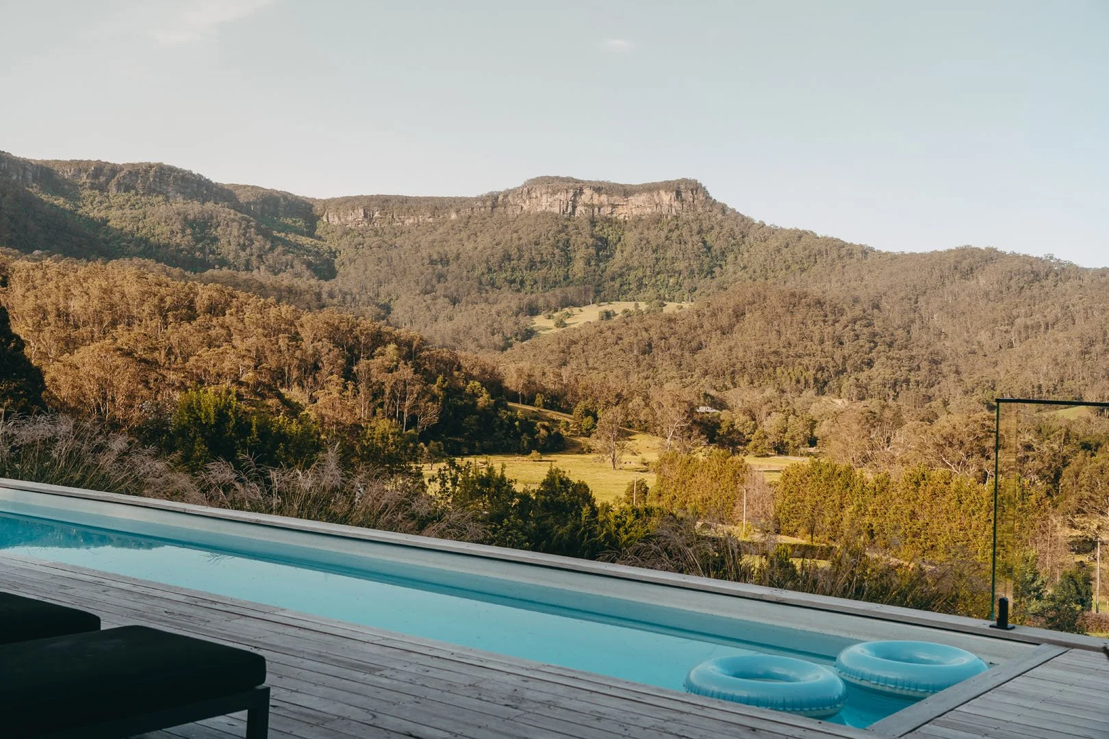 Outdoor pool with mountain landscape in the background, wooden deck, black lounge chair, and inflatable pool floaties.
