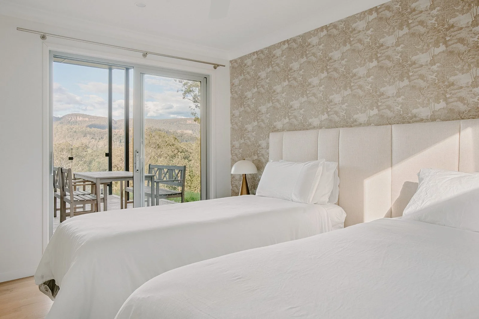 A bedroom with two beds, white bedding, a nightstand with a lamp, and a sliding glass door leading to a balcony with outdoor furniture, overlooking a scenic landscape of hills and trees.