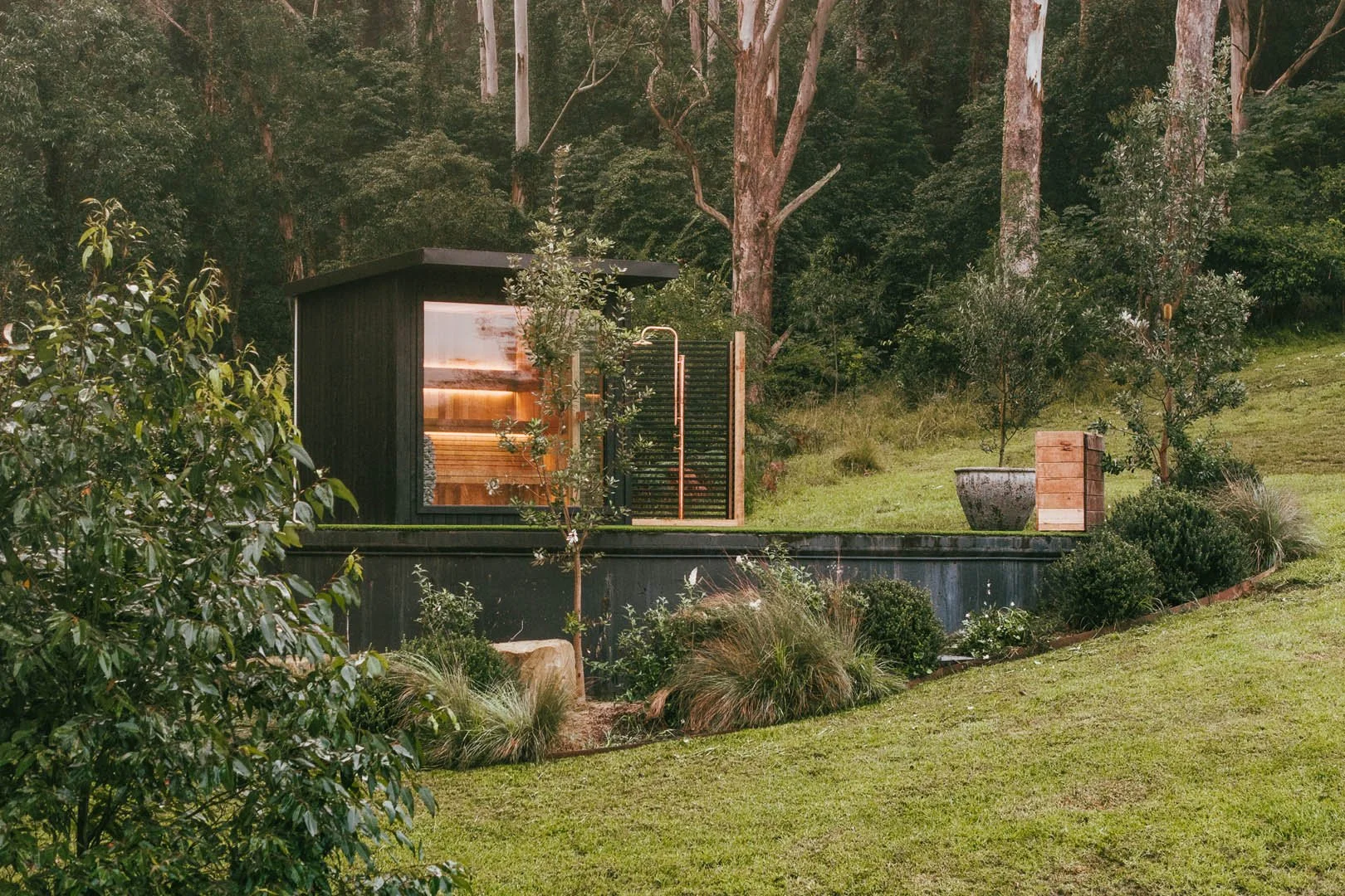 A modern black garden shed with a large glass window, set on a grassy lawn surrounded by bushes and small trees, with taller trees and woodland in the background.