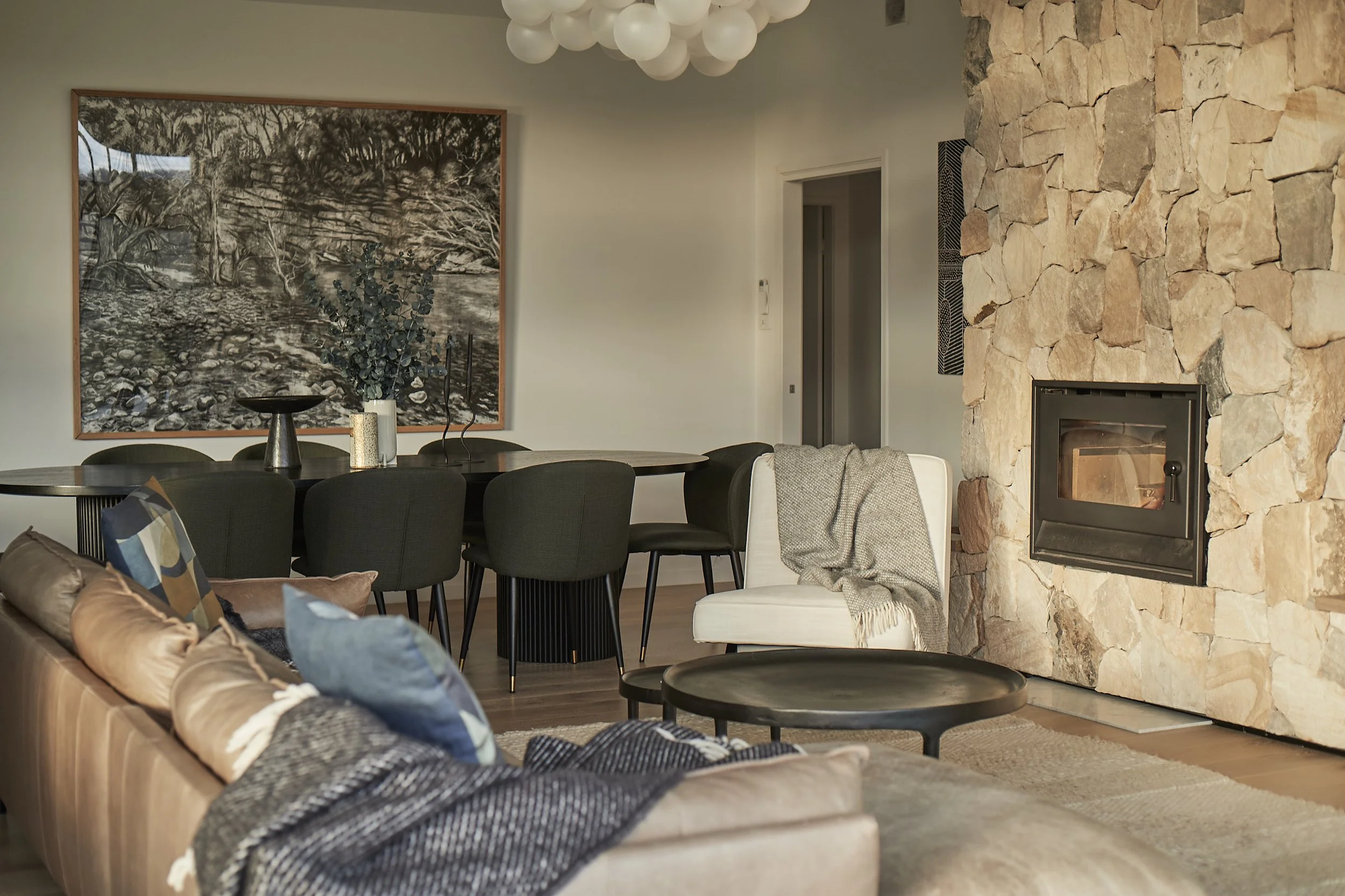 Living room with a beige sofa with colorful cushions, a round black coffee table, a white armchair with a gray throw blanket, a stone fireplace, a dining table with black chairs, wall art, and a chandelier.