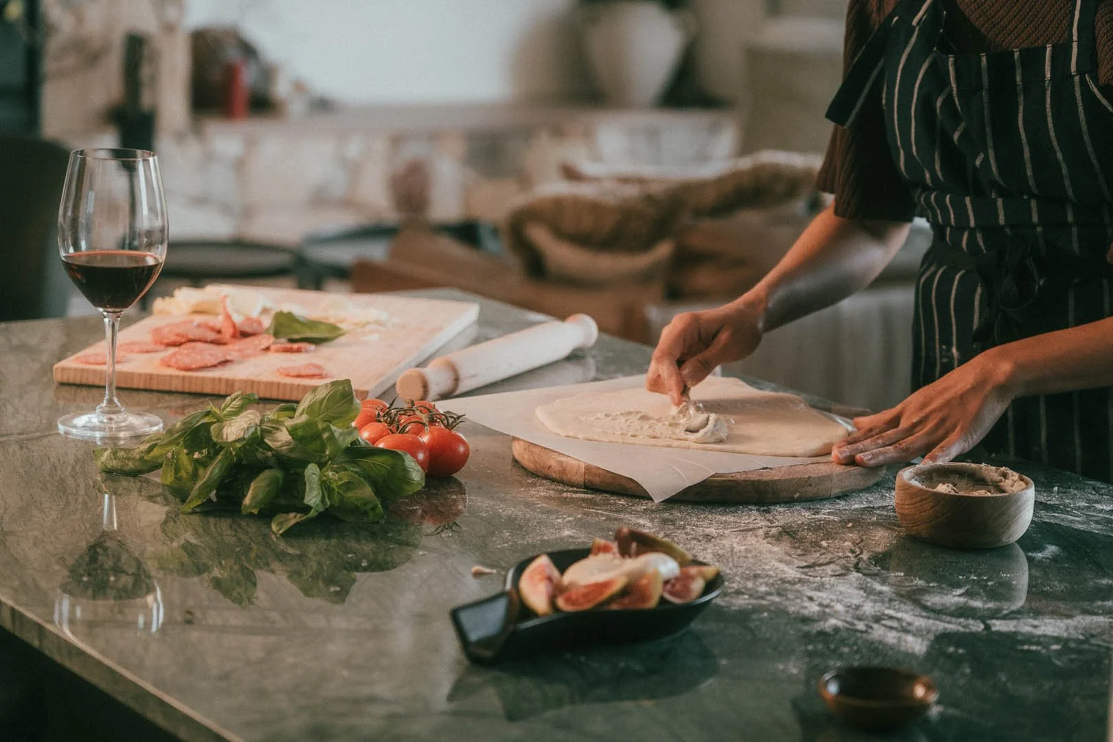 Person preparing pizza dough in a rustic kitchen, with fresh tomatoes, basil, a glass of red wine, and various ingredients on the marble countertop.