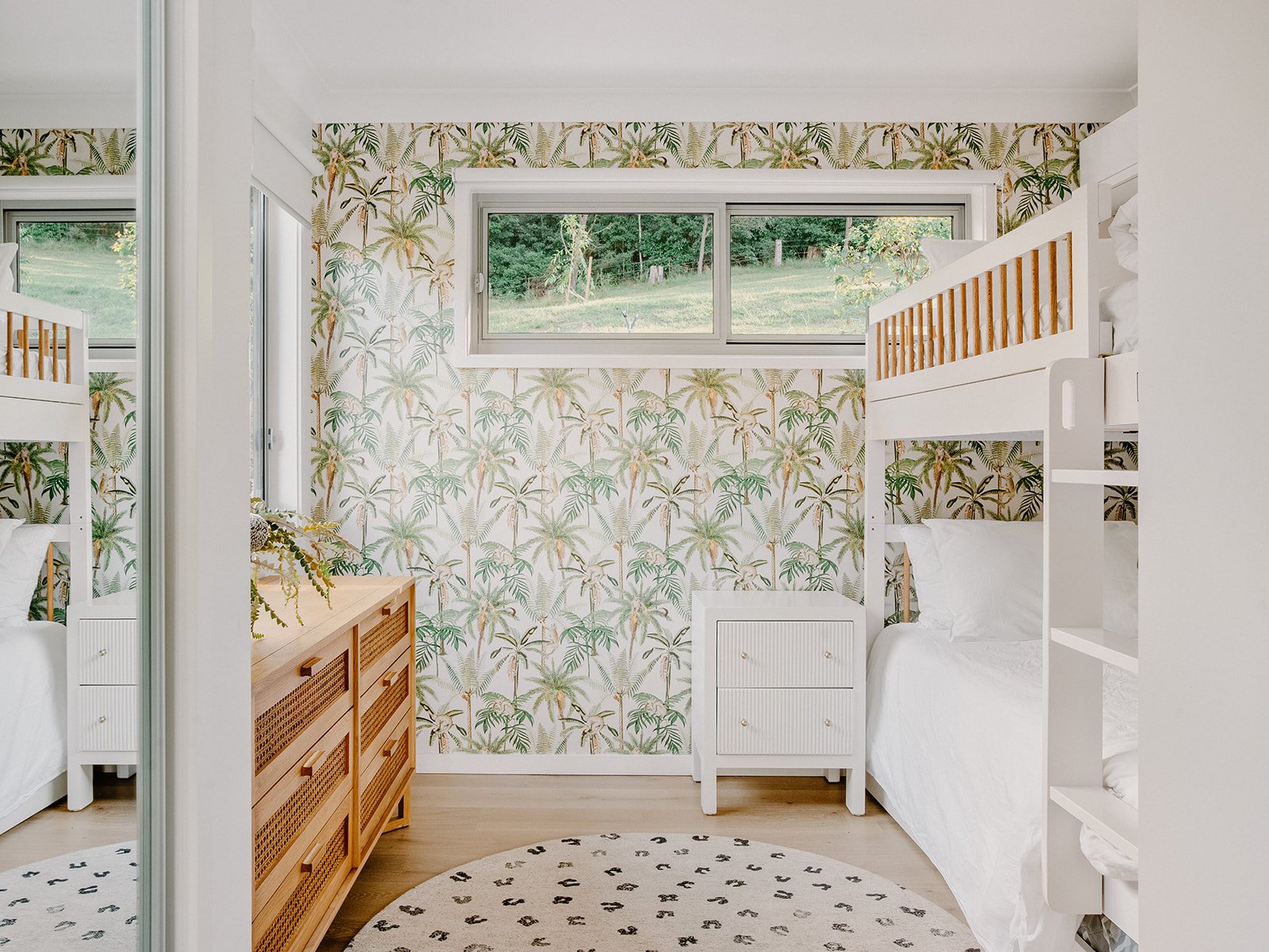 A bedroom with a tropical leaf and palm tree patterned wallpaper, two white bunk beds, a wooden dresser, a white nightstand, and a large window overlooking a grassy yard with trees.
