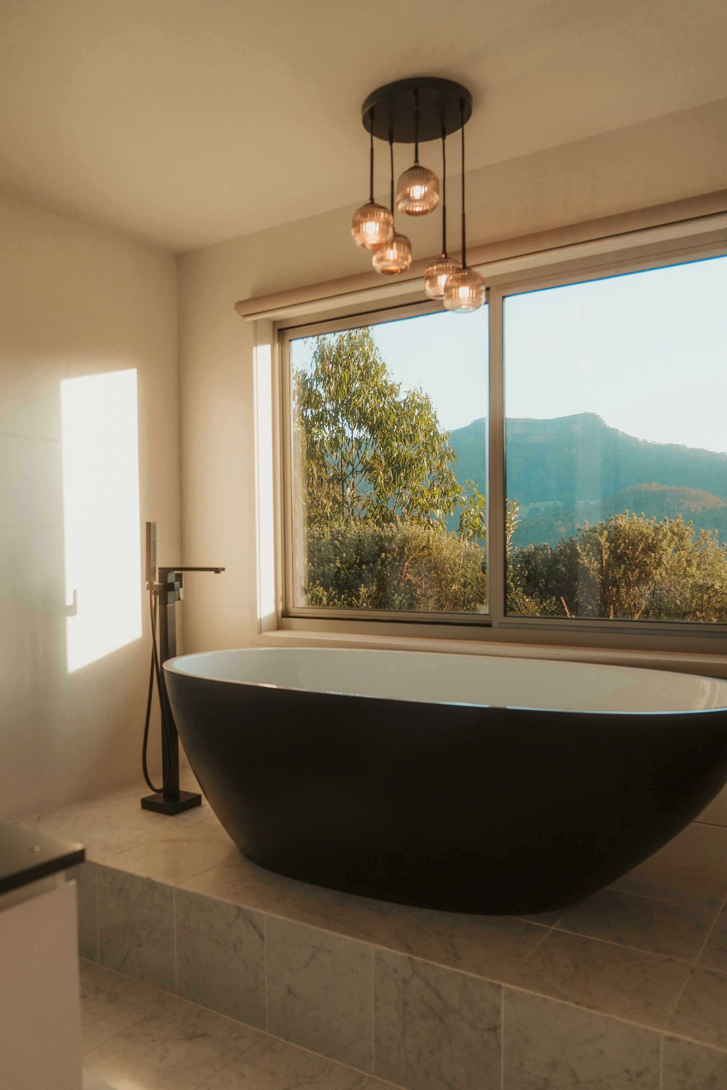 Modern bathroom with a black freestanding bathtub, a large window showing a view of trees and mountains, a ceiling light fixture with multiple bulbs, and a floor-mounted faucet.