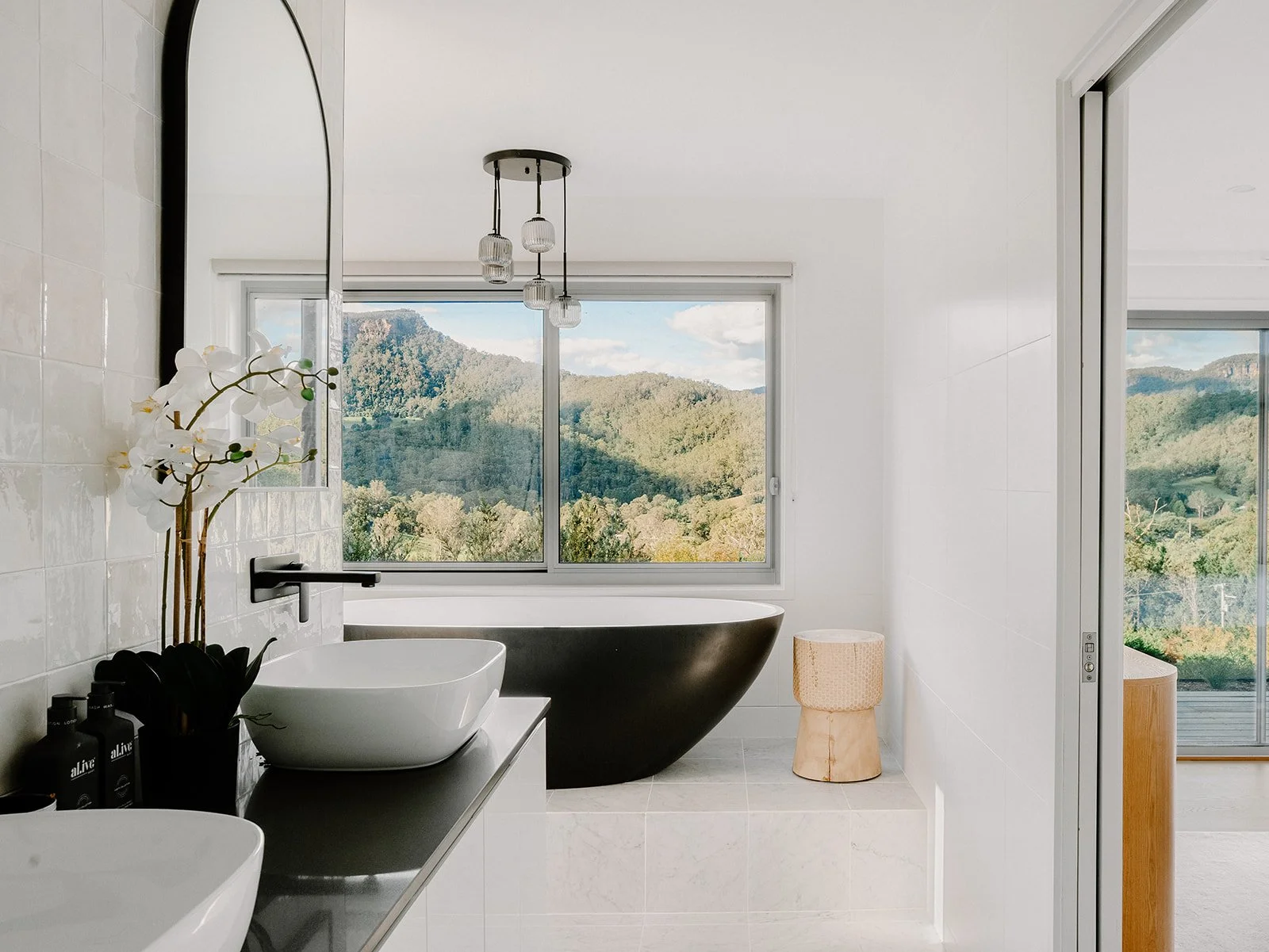 Modern bathroom with a mountain view, black bathtub, white sinks, orchid plant, wooden stool, hanging light fixtures, and sliding glass door to a balcony.
