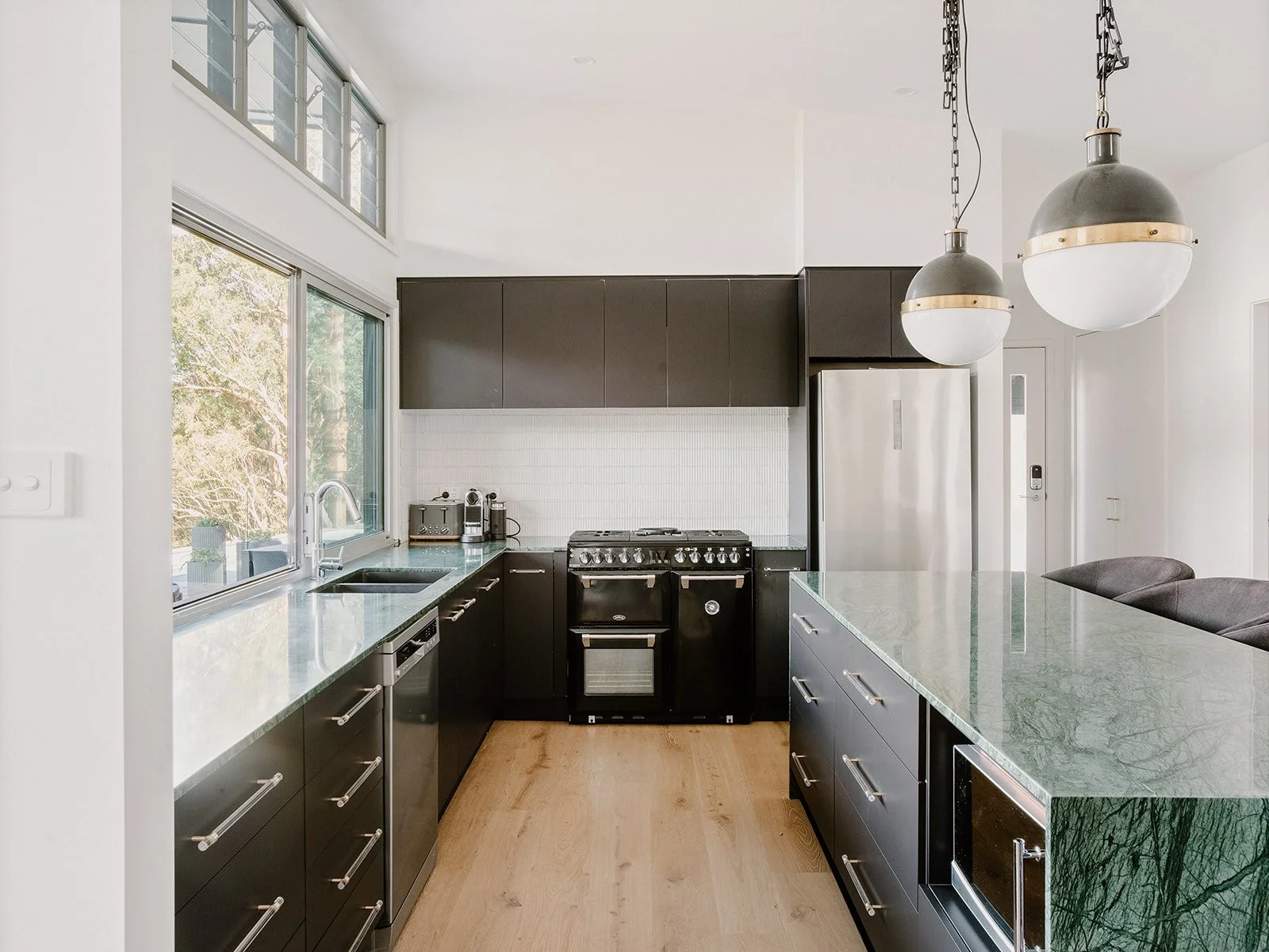 Modern kitchen with black cabinetry, stainless steel appliances, a large window over the sink, green marble countertop on a kitchen island, two hanging pendant lights, and wood flooring.