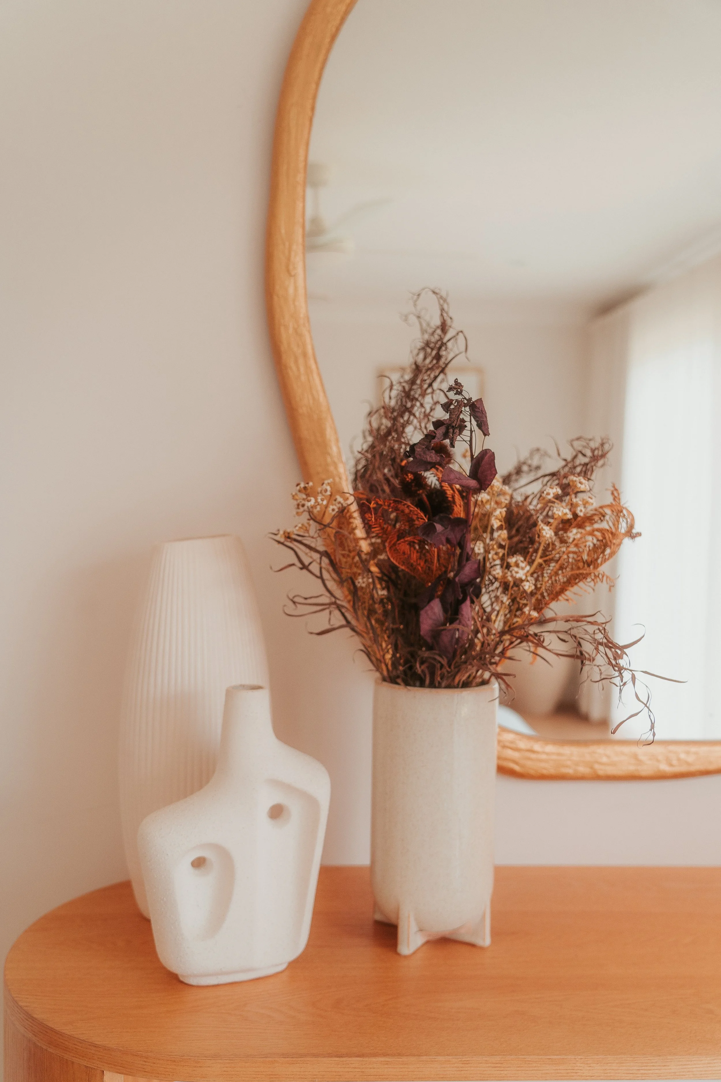 Decorative scene with a dried flower arrangement in a tall, beige vase on a wooden table, with a contemporary white ceramic sculpture and a textured white lamp, reflected in a large, curved mirror on the wall.