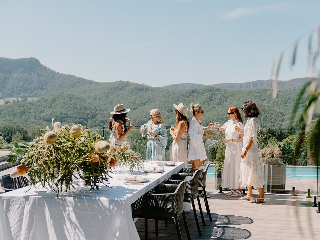 A group of women dressed in white and pastel dresses standing on a deck near a swimming pool, holding drinks and socializing with a scenic hilly landscape in the background on a sunny day.