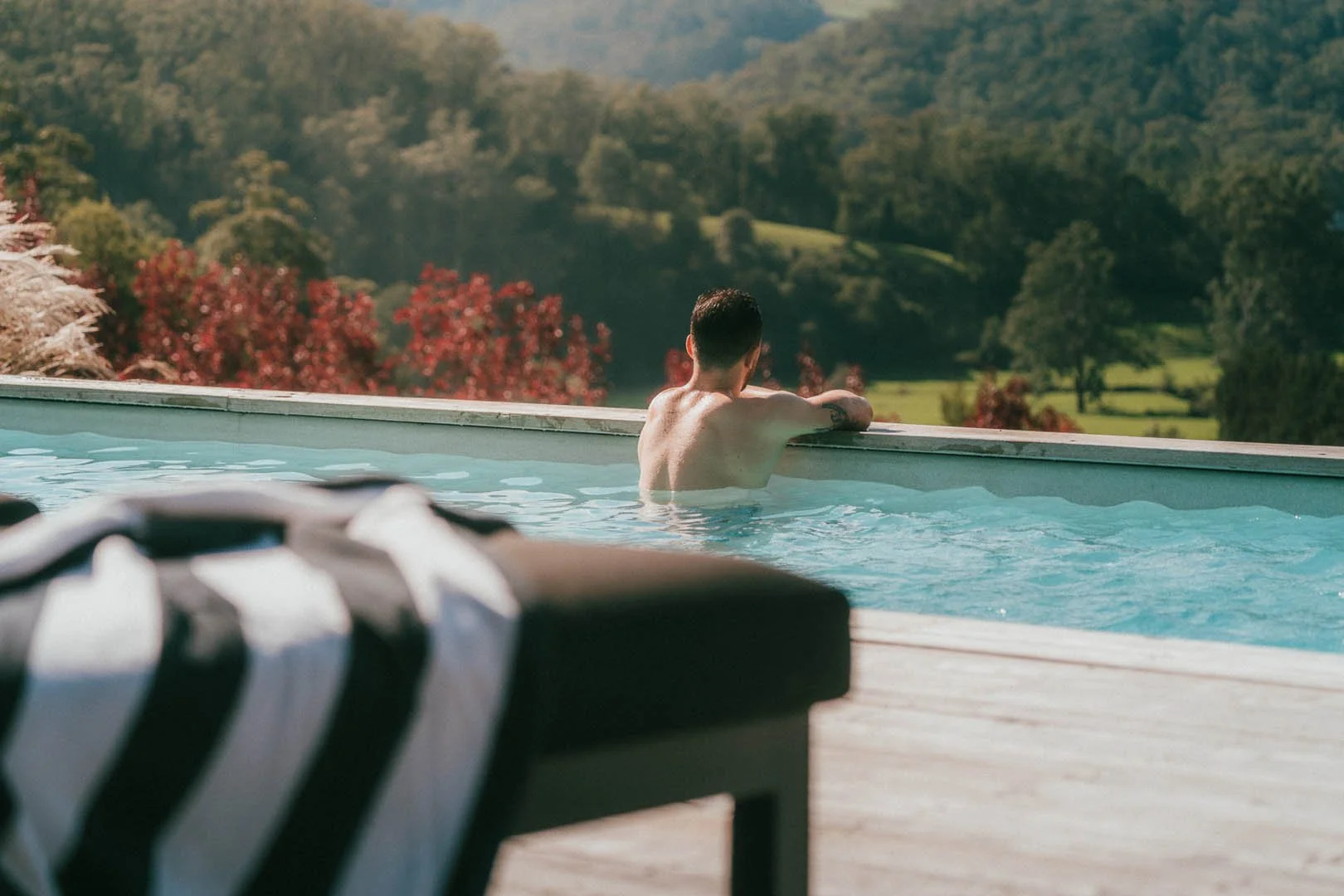 A person relaxing in an outdoor pool, looking at a scenic view of green hills, trees with red foliage, and a bright sky.