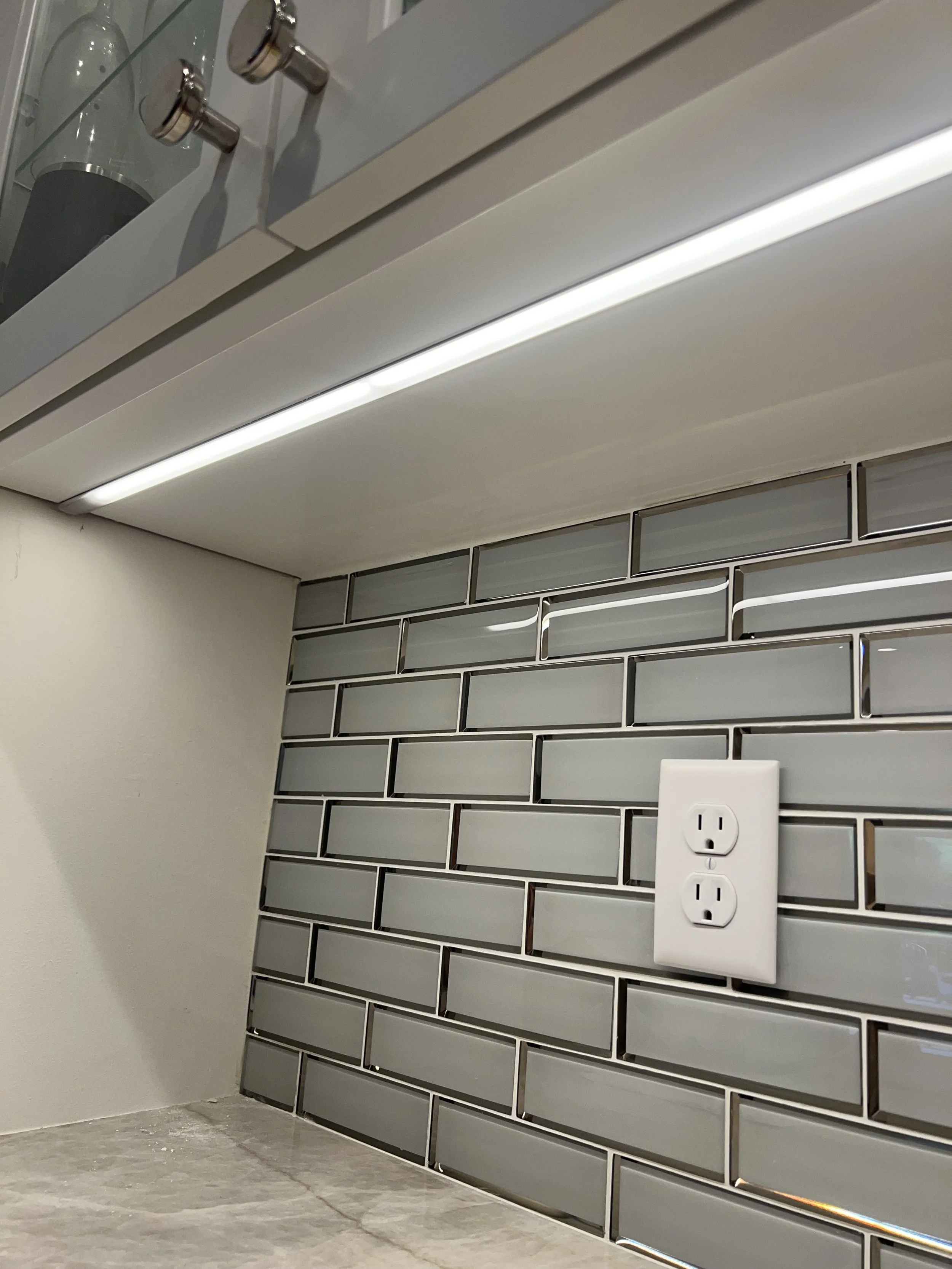 Under-cabinet LED lighting and white electrical outlet installed above a kitchen countertop with gray subway tile backsplash – installed by Walter Electrical Contractors.