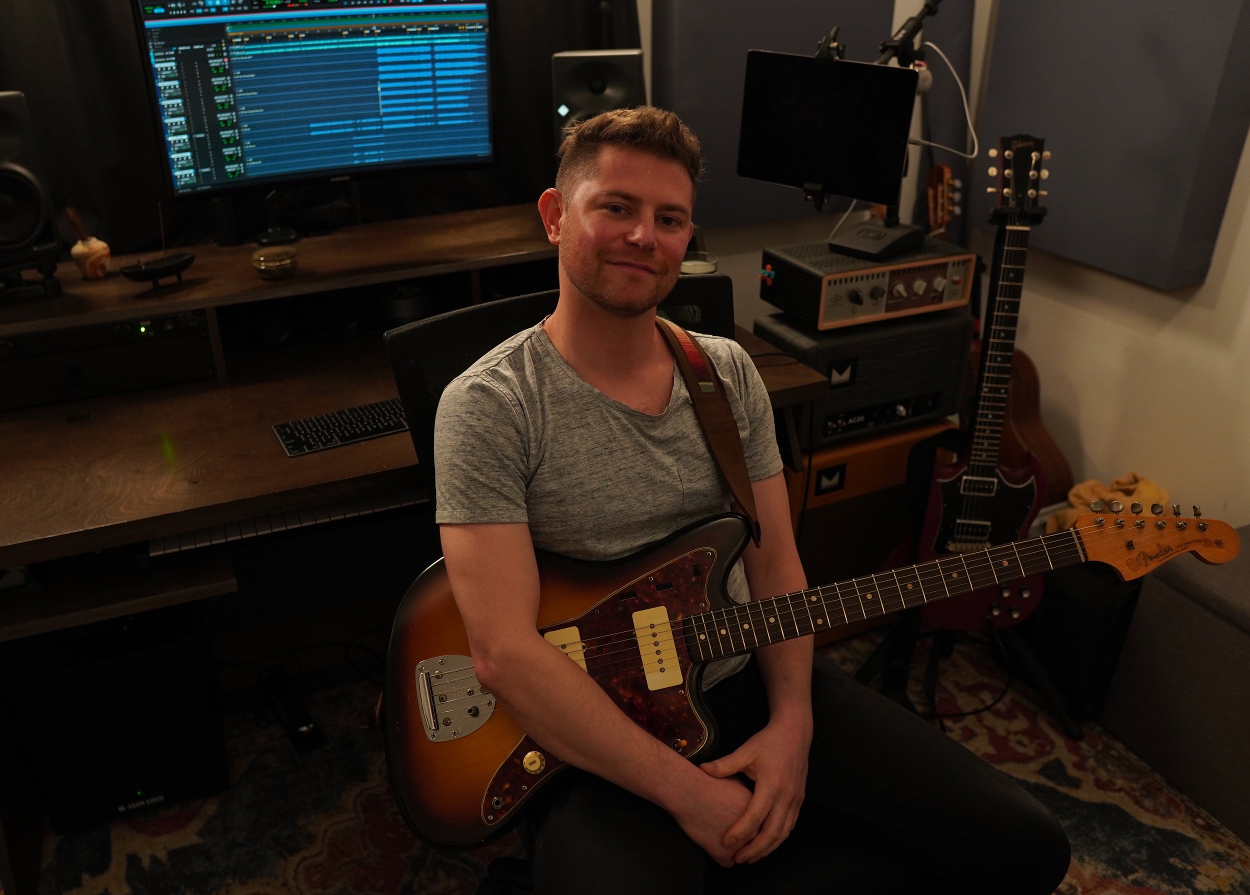 A man sitting in a music studio holding an electric guitar, with music production equipment including a computer monitor, speakers, a keyboard, and guitars in the background.