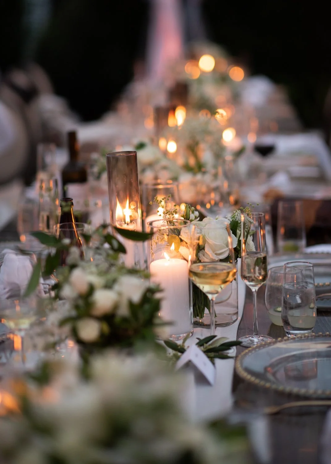 A decorated outdoor dining table illuminated by candles and string lights, adorned with white flowers and glassware.
