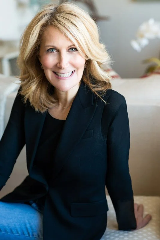 A portrait of Elyse from EAF fine papers. She is a  smiling woman with blonde hair wearing a black blazer sitting on a beige couch in a well-lit room.