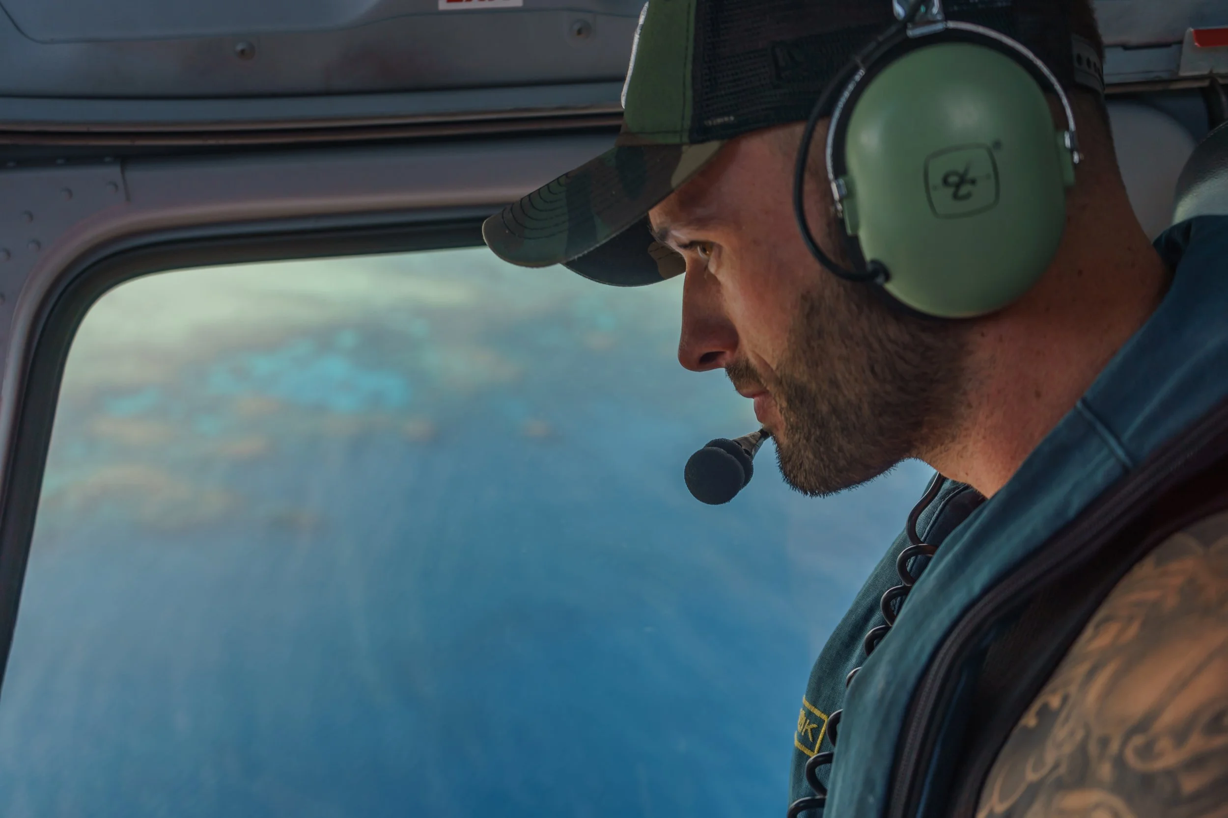 A man wearing a camo cap, green headset, and military uniform flying a helicopter over the ocean.